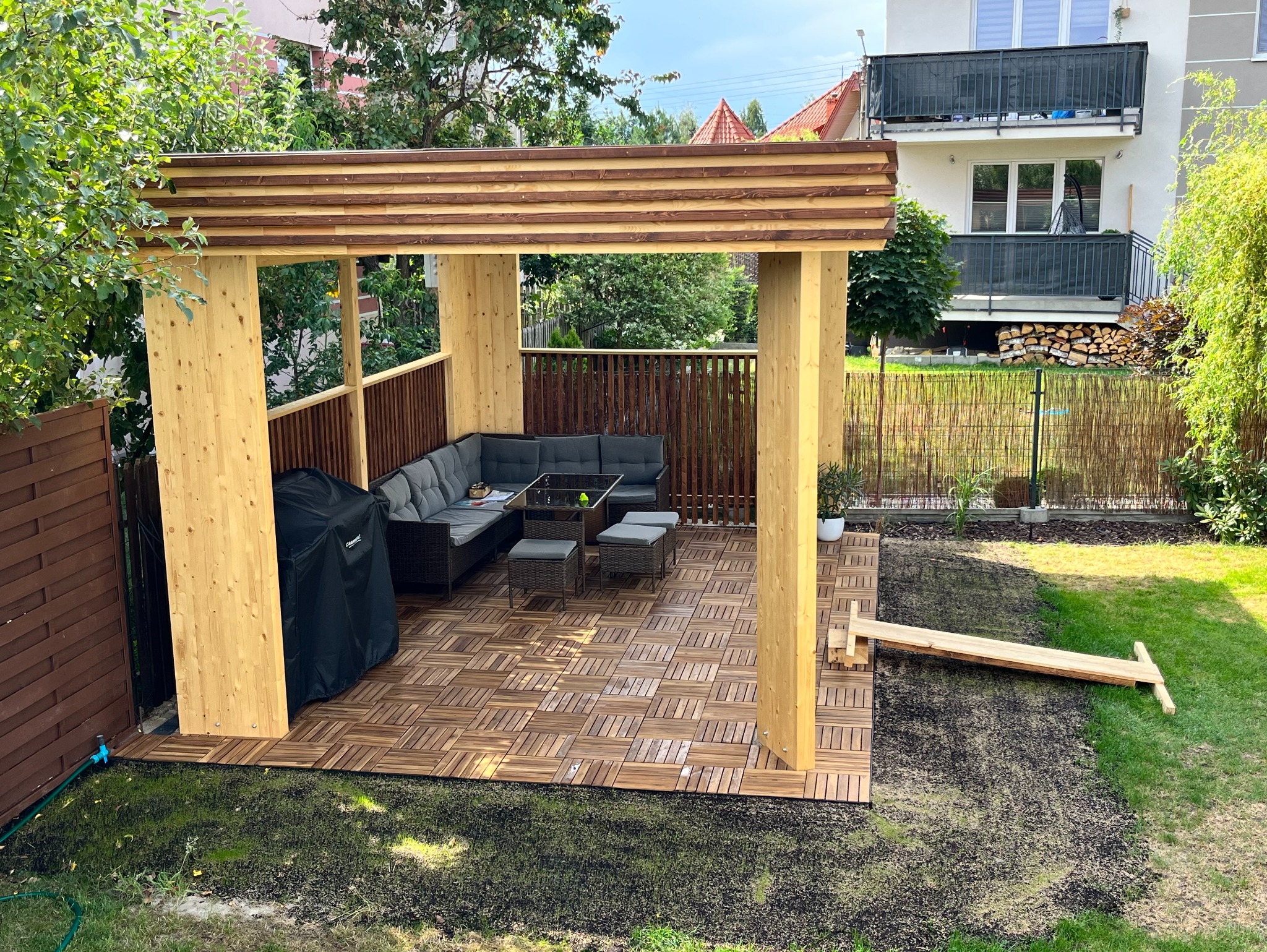 Drewniana altana z zadaszeniem lamelowym, wyposażona w meble ogrodowe z szarego rattanu, grill pod pokrowcem i drewniany podest z kwadratowych płytek na trawniku.