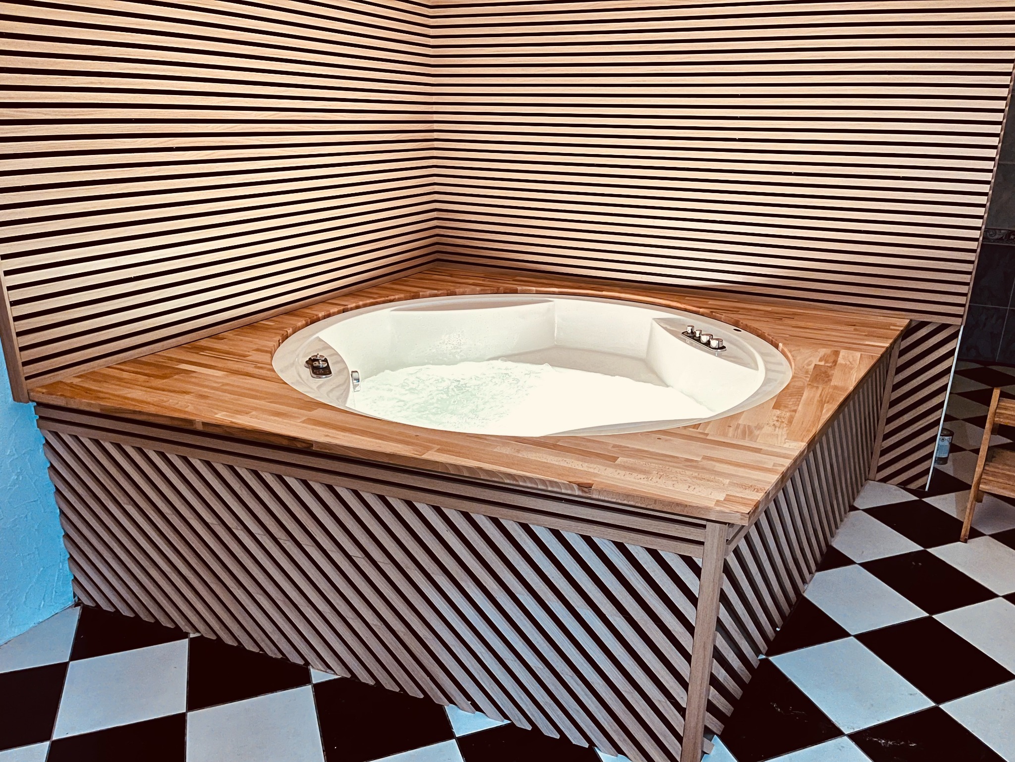 Nowoczesna wanna z hydromasażem, otoczona drewnianą zabudową z listew, na podłodze czarno-biała szachownica, ściany wykończone poziomymi panelami drewnianymi.