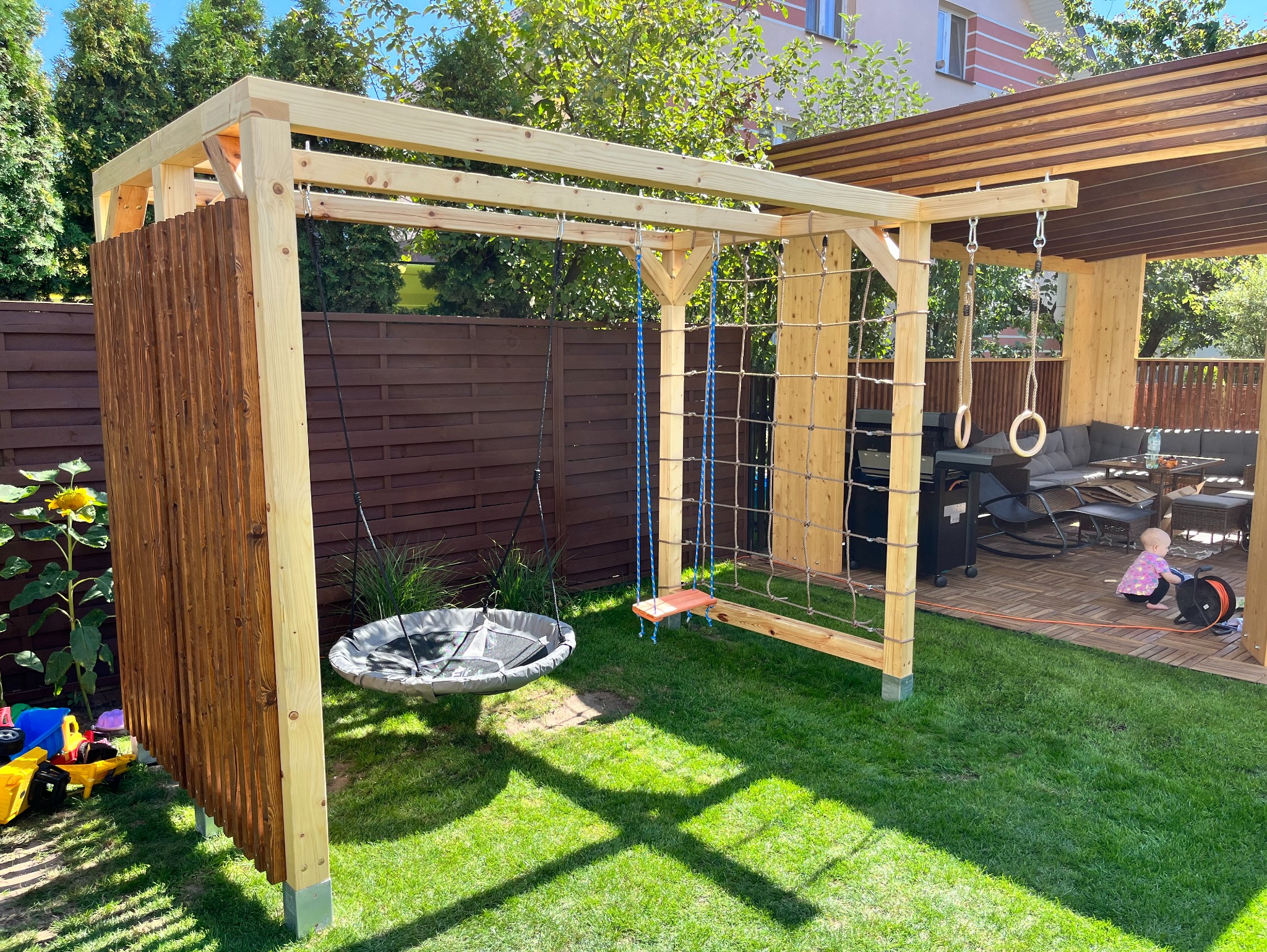 Drewniana konstrukcja placu zabaw w ogrodzie z huśtawkami, siatką do wspinaczki i drewnianym panelem, na trawniku zabawki i dziecko bawiące się na tarasie.