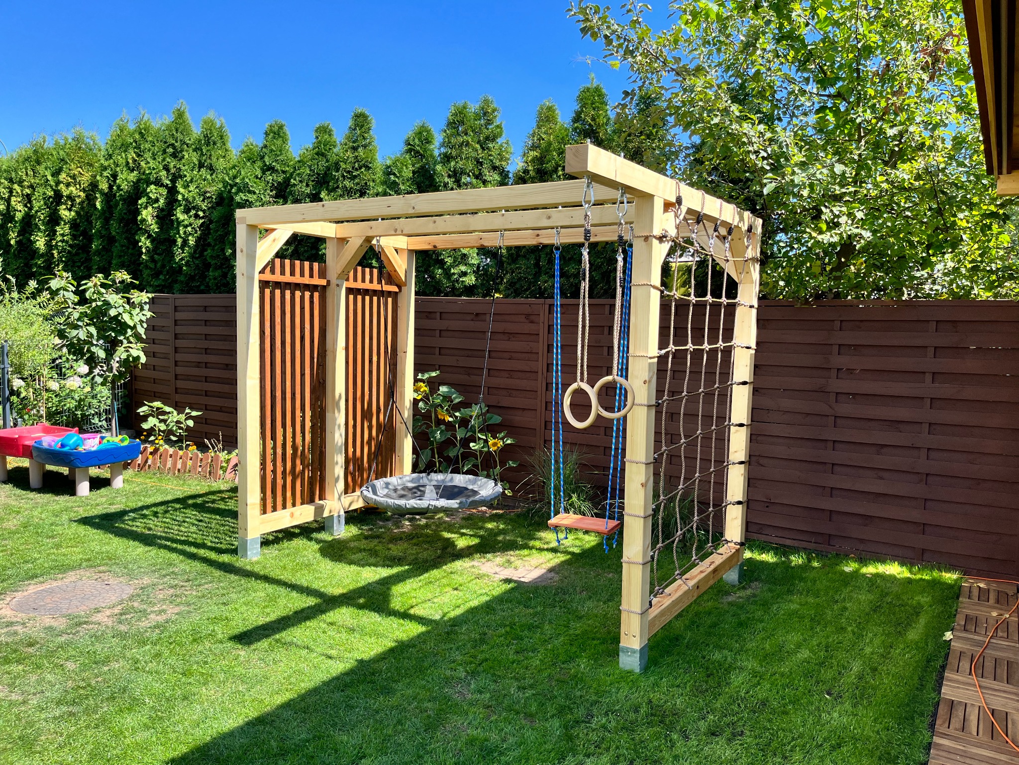 Drewniana konstrukcja placu zabaw w ogrodzie z huśtawkami, drabinką sznurową i ścianką z pionowych listew, otoczona zielenią i brązowym płotem.