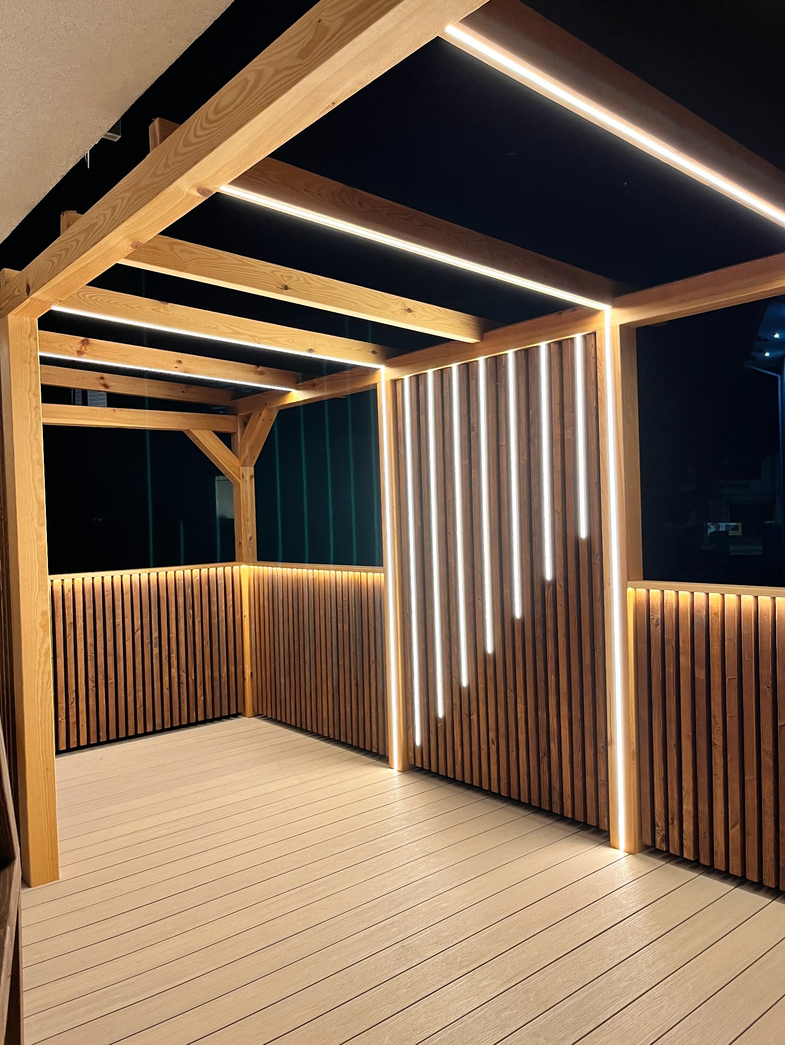 Drewniana altana z nowoczesnym oświetleniem LED, pionowe panele z lamelami i deski podłogowe w ciepłych odcieniach brązu, oświetlona od góry i z boków, nocne ujęcie.