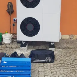 Biała pompa ciepła marki Saunier Duval zamontowana na zewnątrz budynku z pomarańczową elewacją, obok widoczna ozdobna studzienka, a na pierwszym planie niebieska skrzynka z narzędziami i torba...