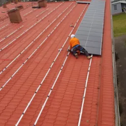 Instalator w pomarańczowej koszuli i kasku montuje panele fotowoltaiczne na czerwonym dachu z metalowej dachówki, widoczne aluminiowe szyny montażowe i przewody elektryczne.