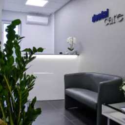 Wnętrze poczekalni: szara sofa, biała recepcja z podświetleniem, roślina doniczkowa w kwadratowej donicy, logo na ścianie, szare płytki podłogowe.