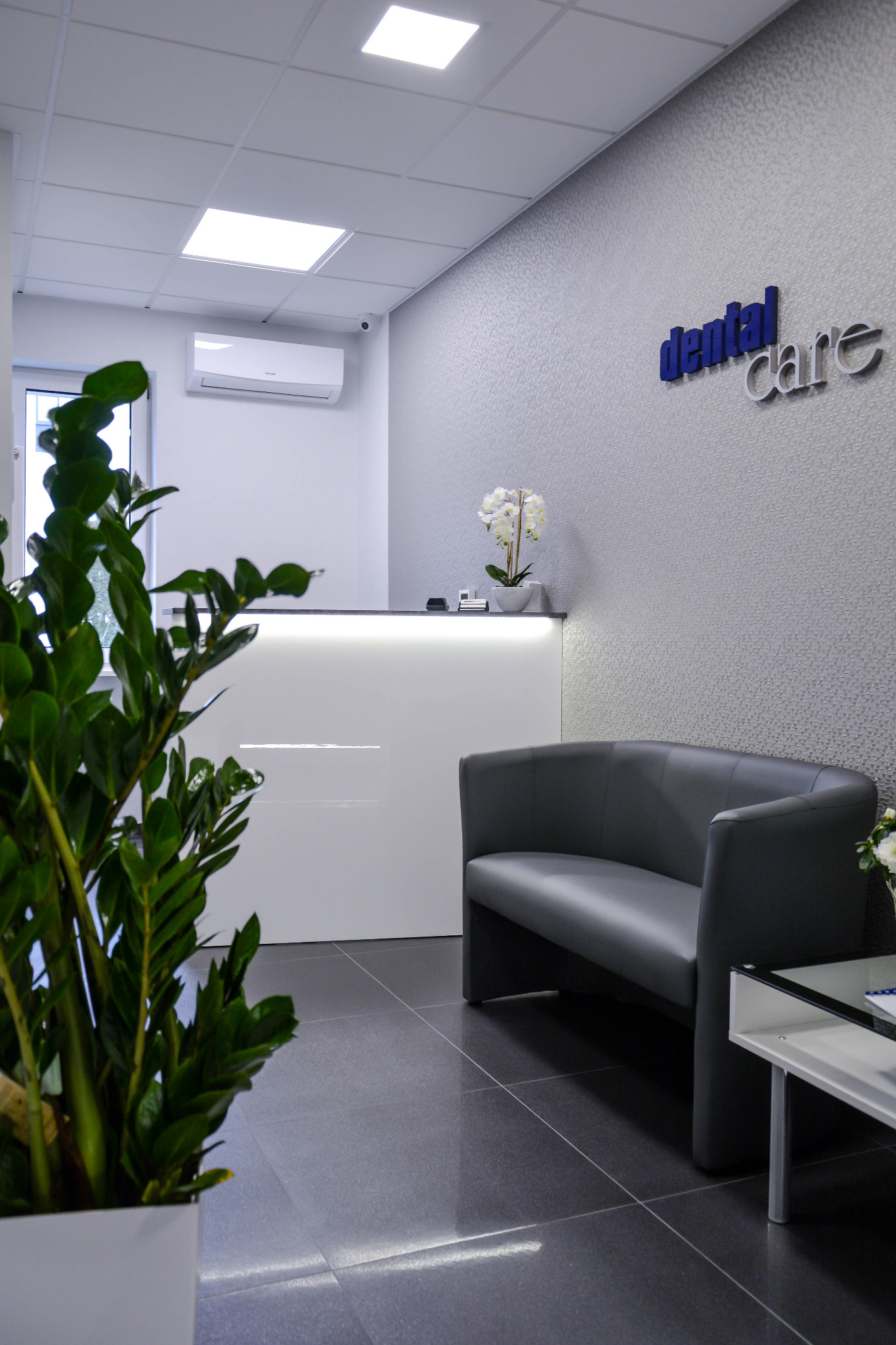 Wnętrze poczekalni: szara sofa, biała recepcja z podświetleniem, roślina doniczkowa w kwadratowej donicy, logo na ścianie, szare płytki podłogowe.