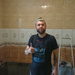 michal papaj - Mężczyzna w roboczym ubraniu, z uniesionym kciukiem, prezentuje częściowo wykafelkowaną łazienkę z beżowo-brązowymi płytkami i widocznym przygotowaniem do dalszych prac wykończeniowych.