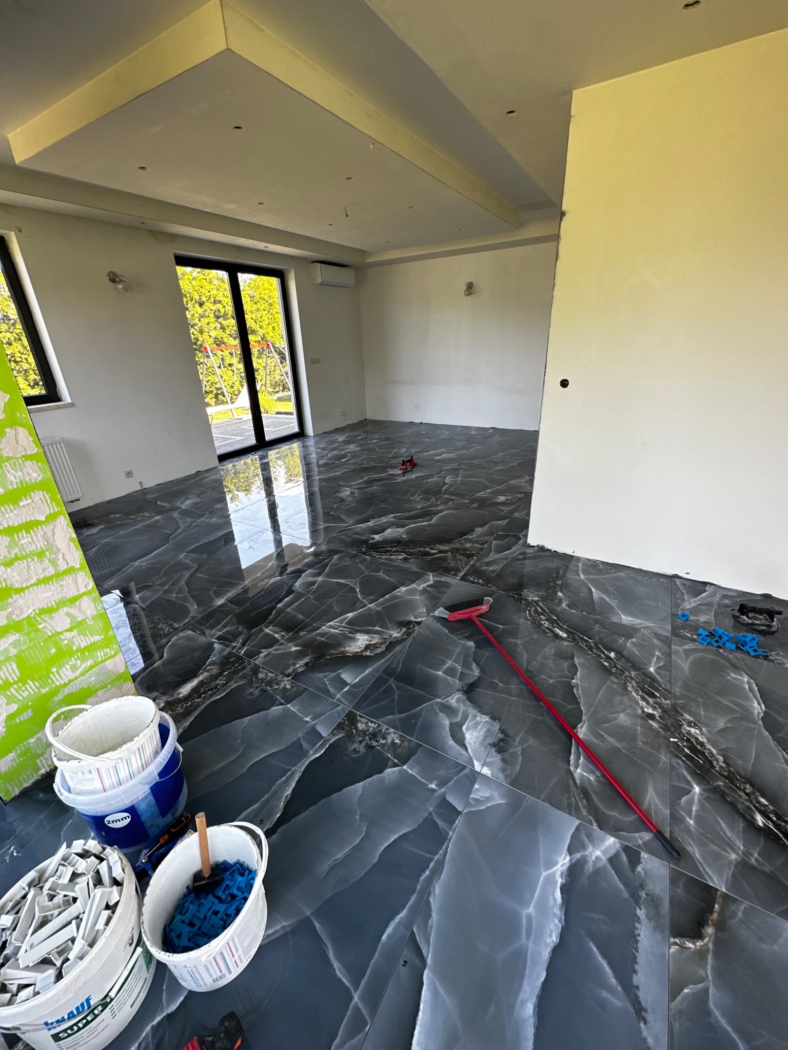 Układanie płytek z efektem marmuru na podłodze w przestronnym, jasnym pomieszczeniu; widoczne narzędzia i materiały budowlane: wiadro z klejem, listwa przypodłogowa, krzyżyki dystansowe, czerwona...