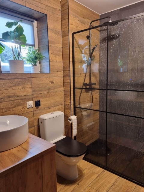 Nowoczesna łazienka z drewnopodobnymi płytkami, kabina prysznicowa typu walk-in z czarnymi profilami, biała umywalka nablatowa na drewnianej szafce, toaleta z czarną deską, rośliny w oknie.