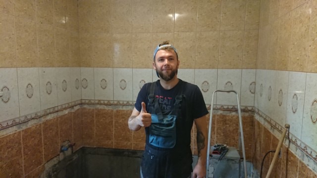 Mężczyzna w roboczym ubraniu, z uniesionym kciukiem, prezentuje częściowo wykafelkowaną łazienkę z beżowo-brązowymi płytkami i widocznym przygotowaniem do dalszych prac wykończeniowych.