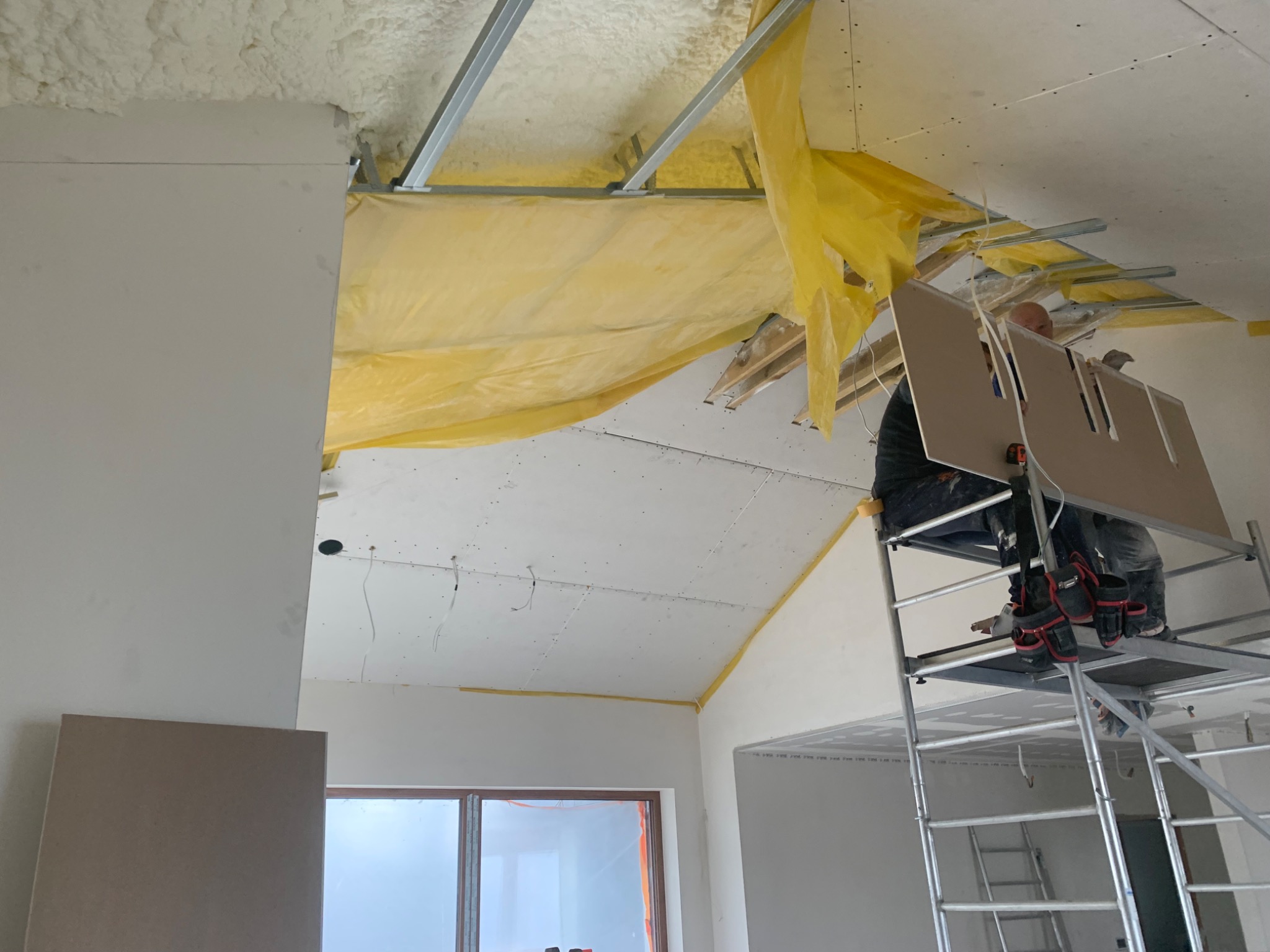 Pracownik na rusztowaniu montuje płytę gipsowo-kartonową na suficie poddasza, widoczne metalowe profile, ocieplenie pianką i folia zabezpieczająca.