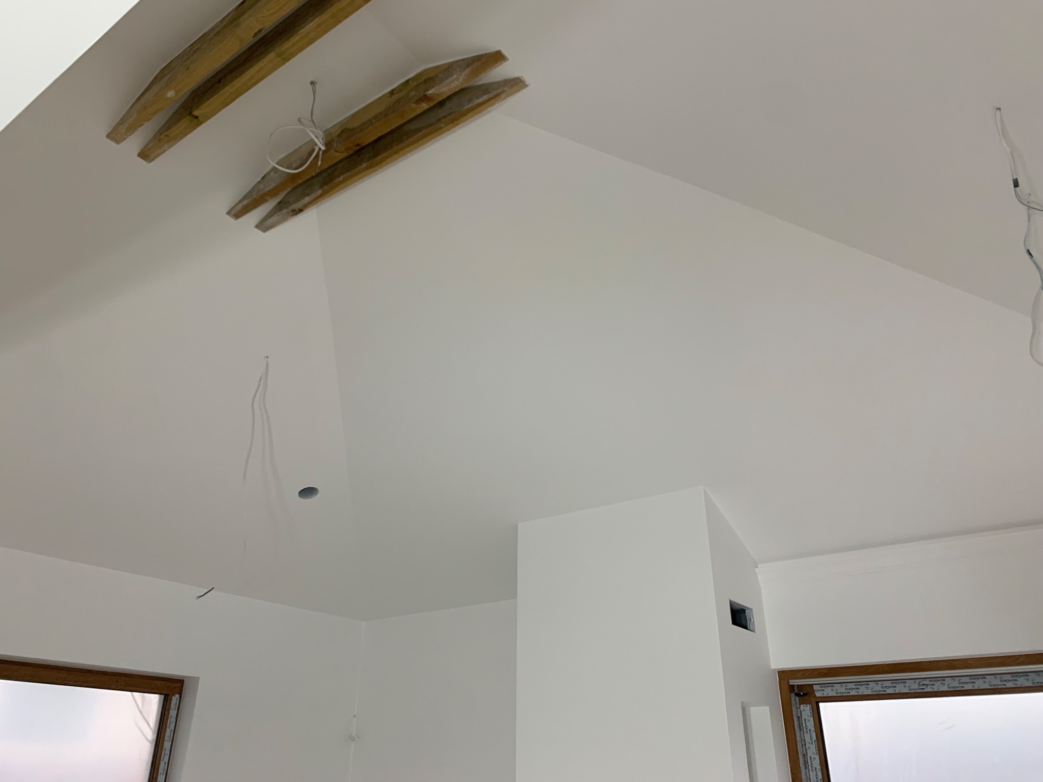 Wnętrze pokoju ze skośnym sufitem po malowaniu na biało, widoczne drewniane belki stropowe, okna z brązowymi ramami i wystające przewody elektryczne.