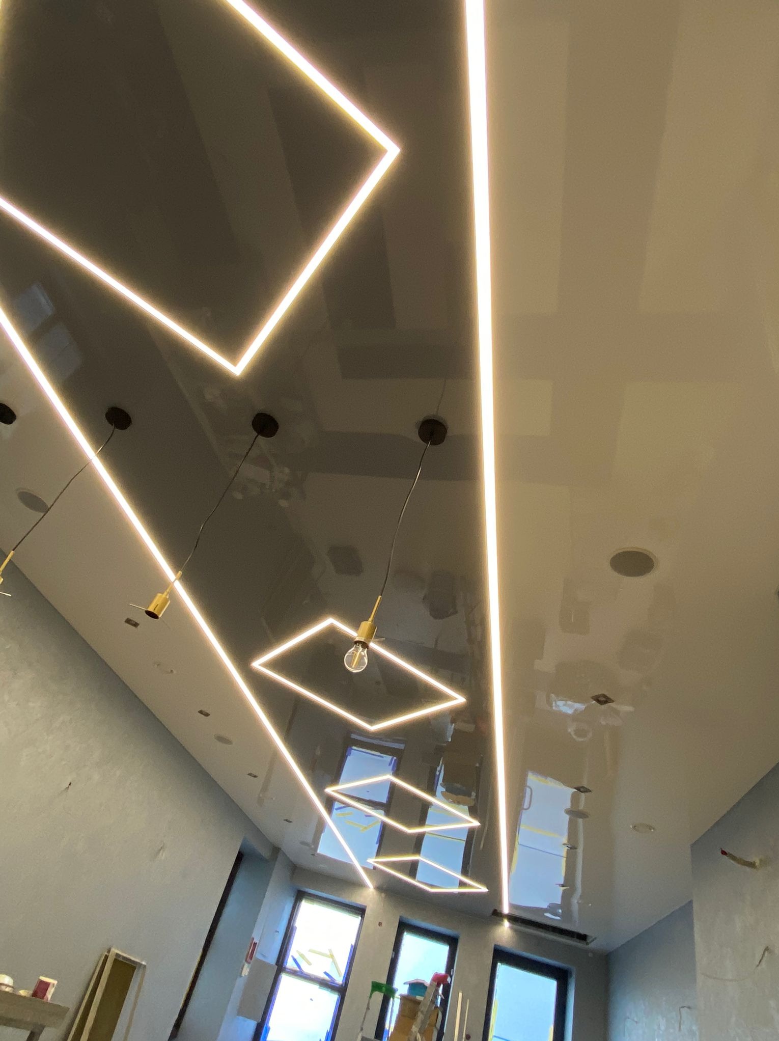 Błyszczący sufit z geometrycznymi, neonowymi lampami i zwisającymi żarówkami, widok z dołu. Nowoczesne oświetlenie w szaro-białym wnętrzu.