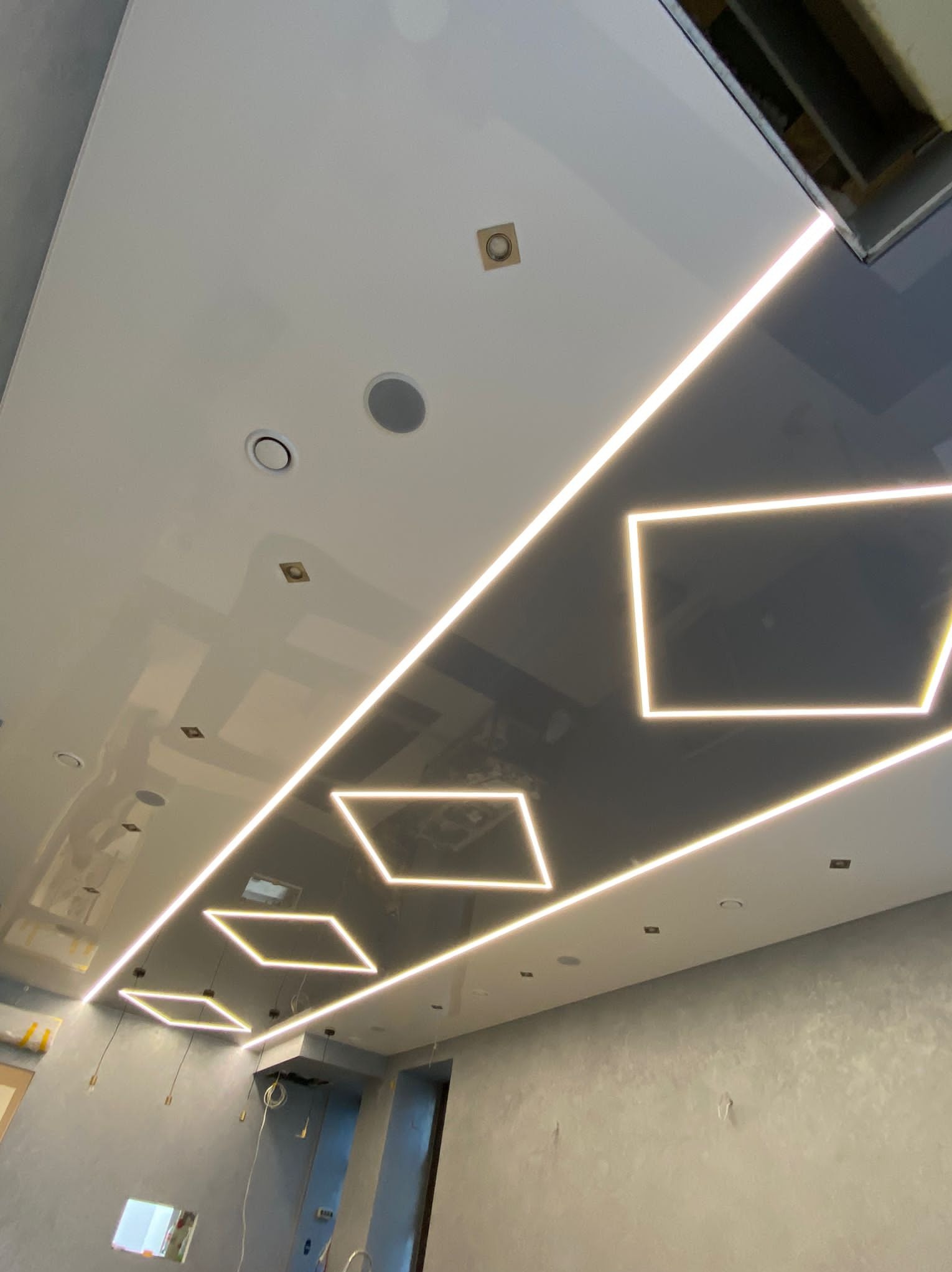 Biało-szary sufit z połyskiem i podświetleniem LED w kształcie rombów i prostokątów. Widoczne głośniki i punkty świetlne. Nowoczesny design.