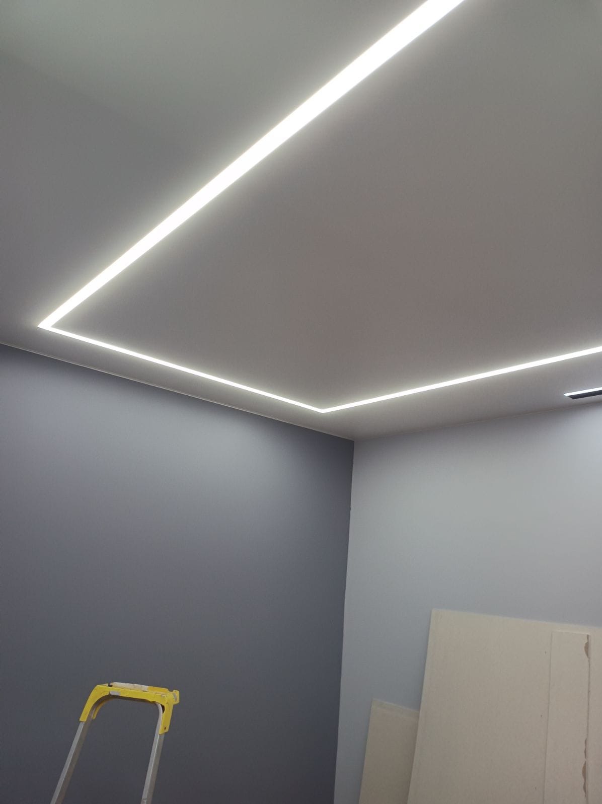 Nowoczesne oświetlenie LED w kształcie prostokąta, wbudowane w jasny sufit. Szara ściana i fragment drabiny malarskiej w dolnej części kadru.