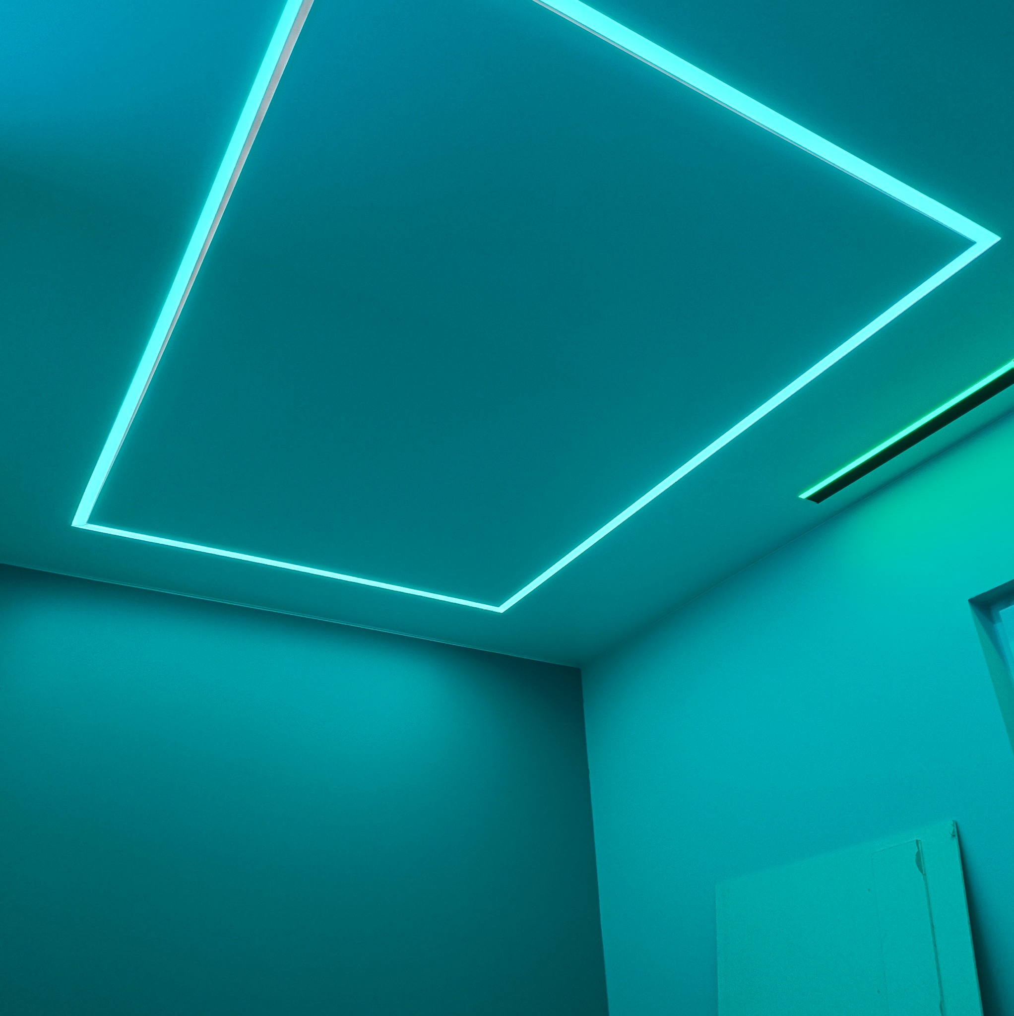 Pokój w odcieniach turkusu z podświetlonym, prostokątnym panelem sufitowym i dodatkowym oświetleniem liniowym, tworzącymi nowoczesny design.