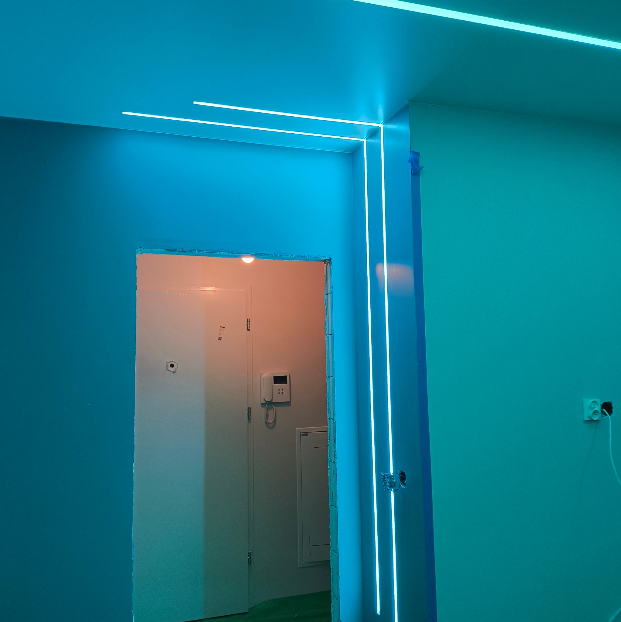 Niebieskie, neonowe oświetlenie LED wzdłuż ścian i sufitu, tworzące nowoczesny design wnętrza. Widoczne surowe wykończenie wokół drzwi i wystające kable.