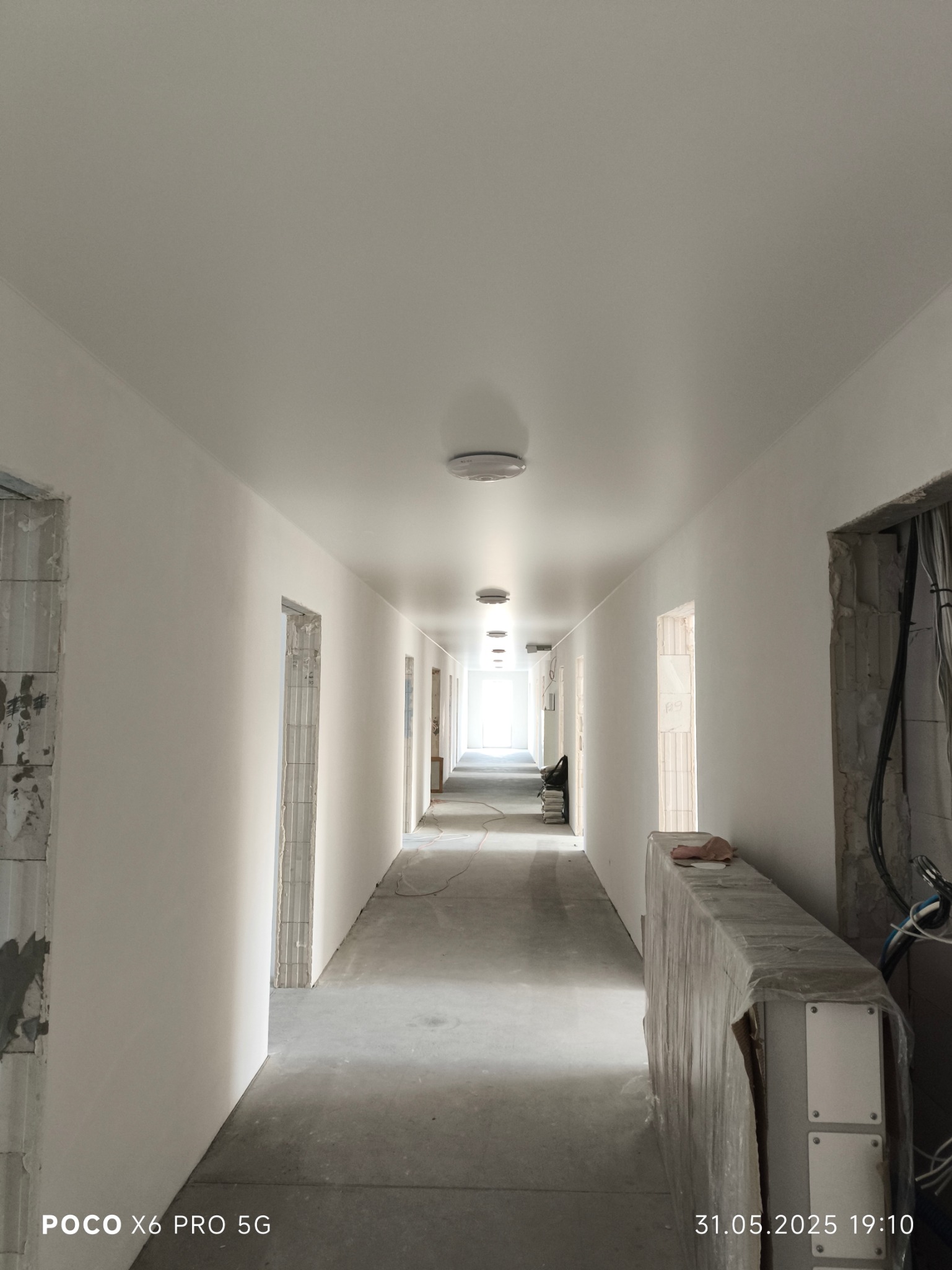 Długi korytarz z nowymi, białymi sufitami i ścianami. Widoczne otwory drzwiowe w trakcie wykończenia. Na podłodze surowy beton.