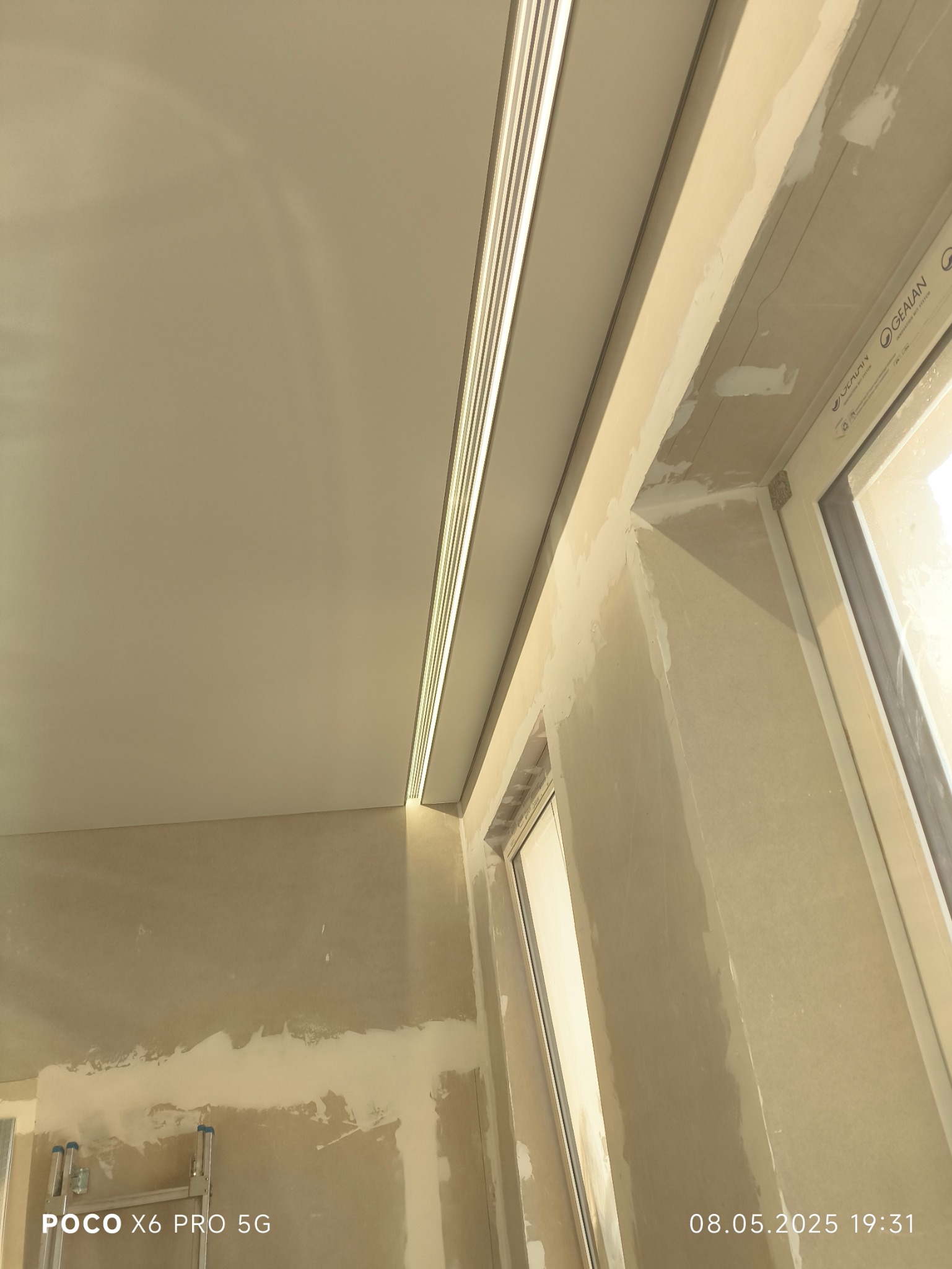 Montaż sufitu napinanego z oświetleniem LED wzdłuż okna. Widoczne niedokończone ściany i listwa sufitowa z oświetleniem. Ujęcie z dołu, ukośne.