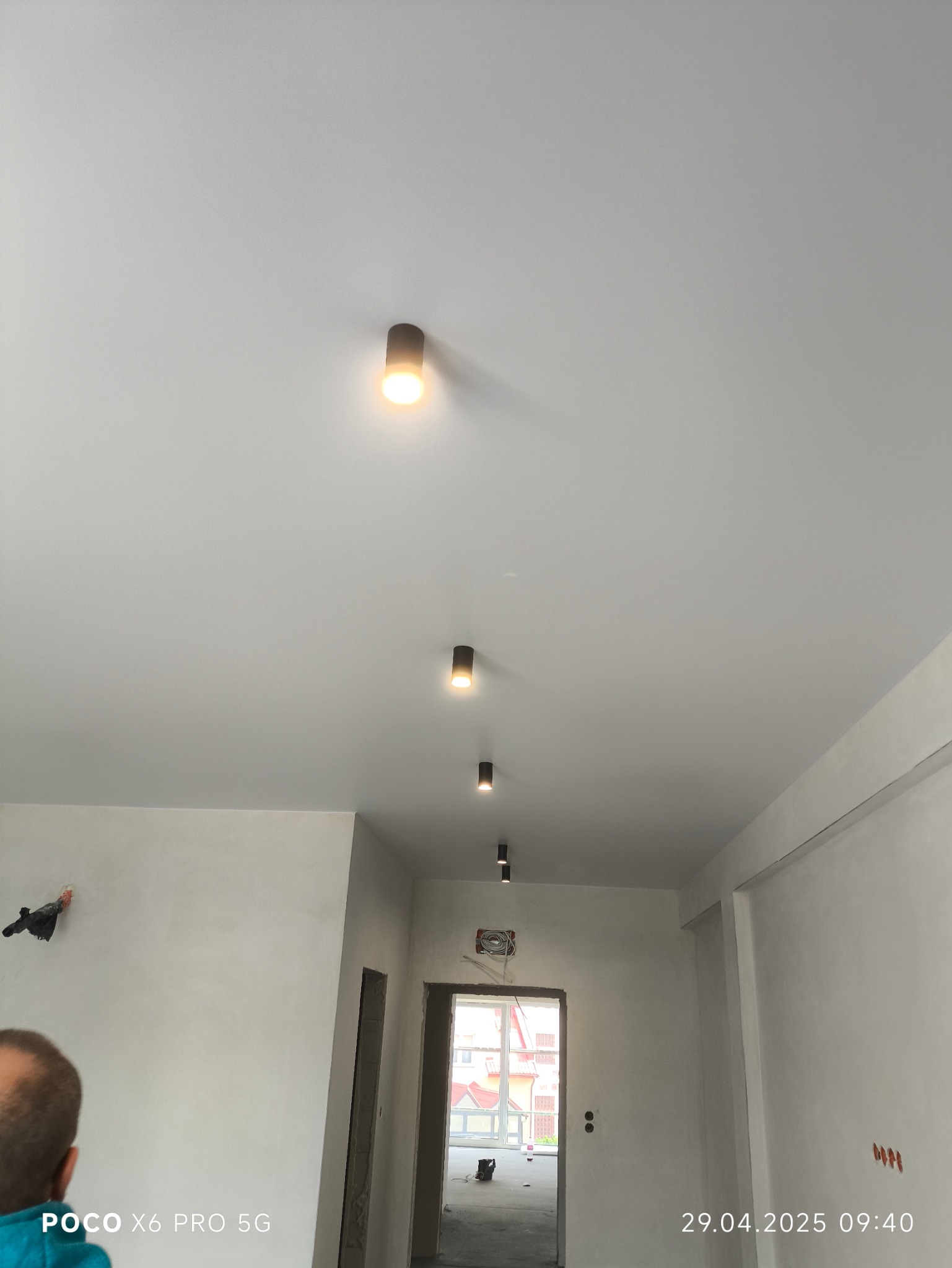 Biały sufit z rzędem czarnych, punktowych lamp sufitowych w długim korytarzu w trakcie wykańczania. Widoczne surowe ściany i przygotowane otwory drzwiowe.