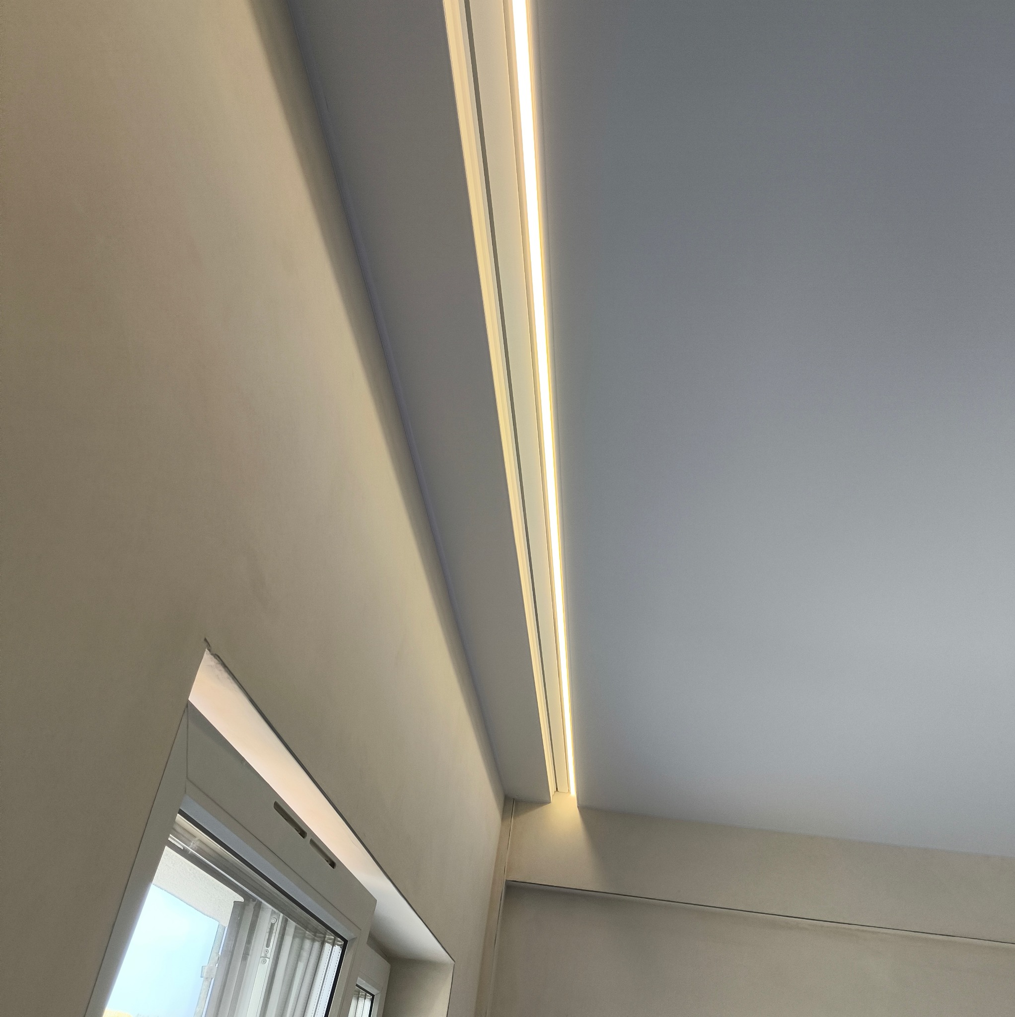 Szary sufit z subtelnym oświetleniem LED wzdłuż krawędzi, widok z dołu, z fragmentem okna po lewej stronie. Nowoczesne wykończenie wnętrza.