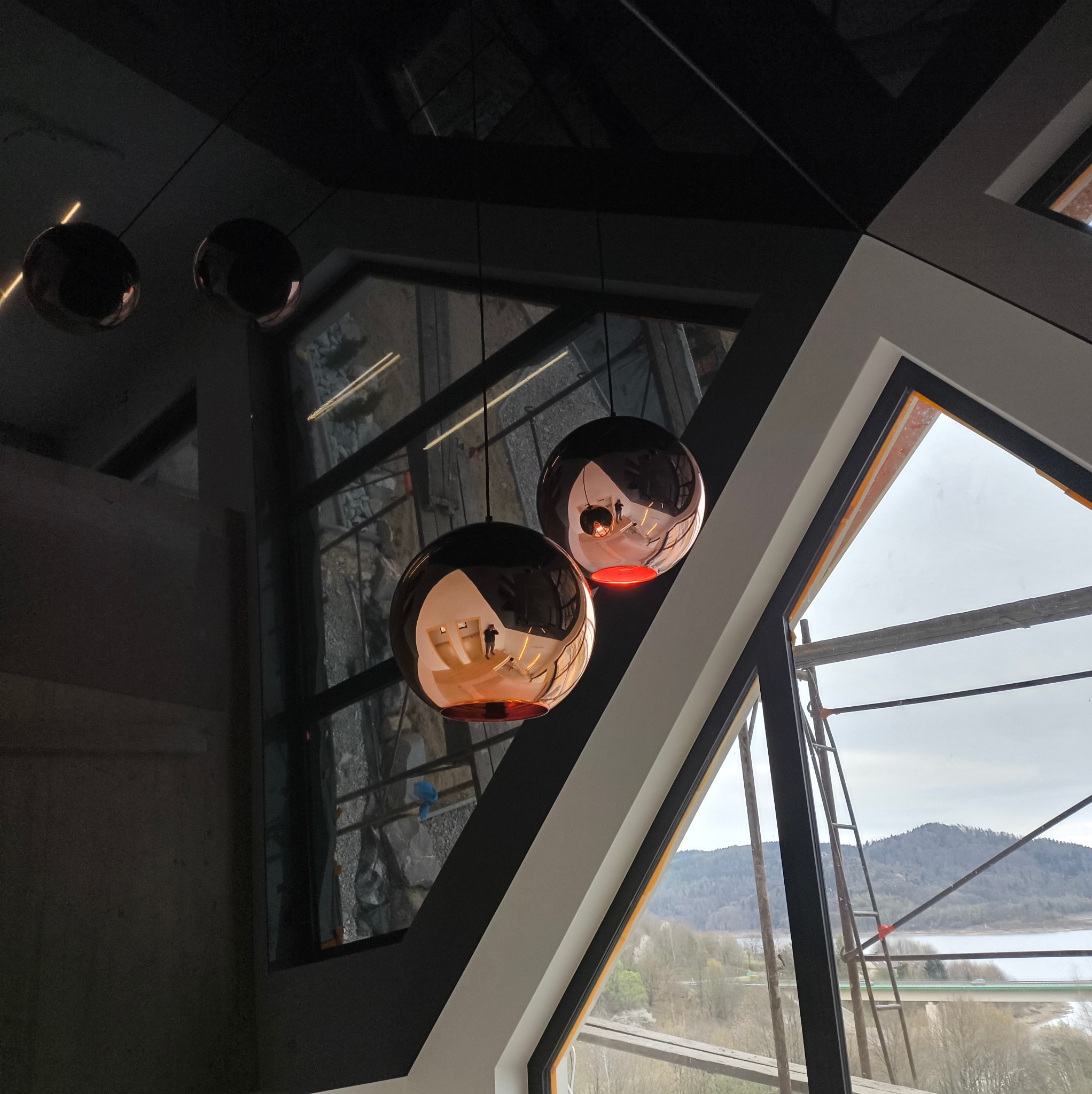 Nowoczesny sufit napinany z połyskliwymi, kulistymi lampami odbijającymi wnętrze. Widok na zewnątrz przez okno z rusztowaniem.