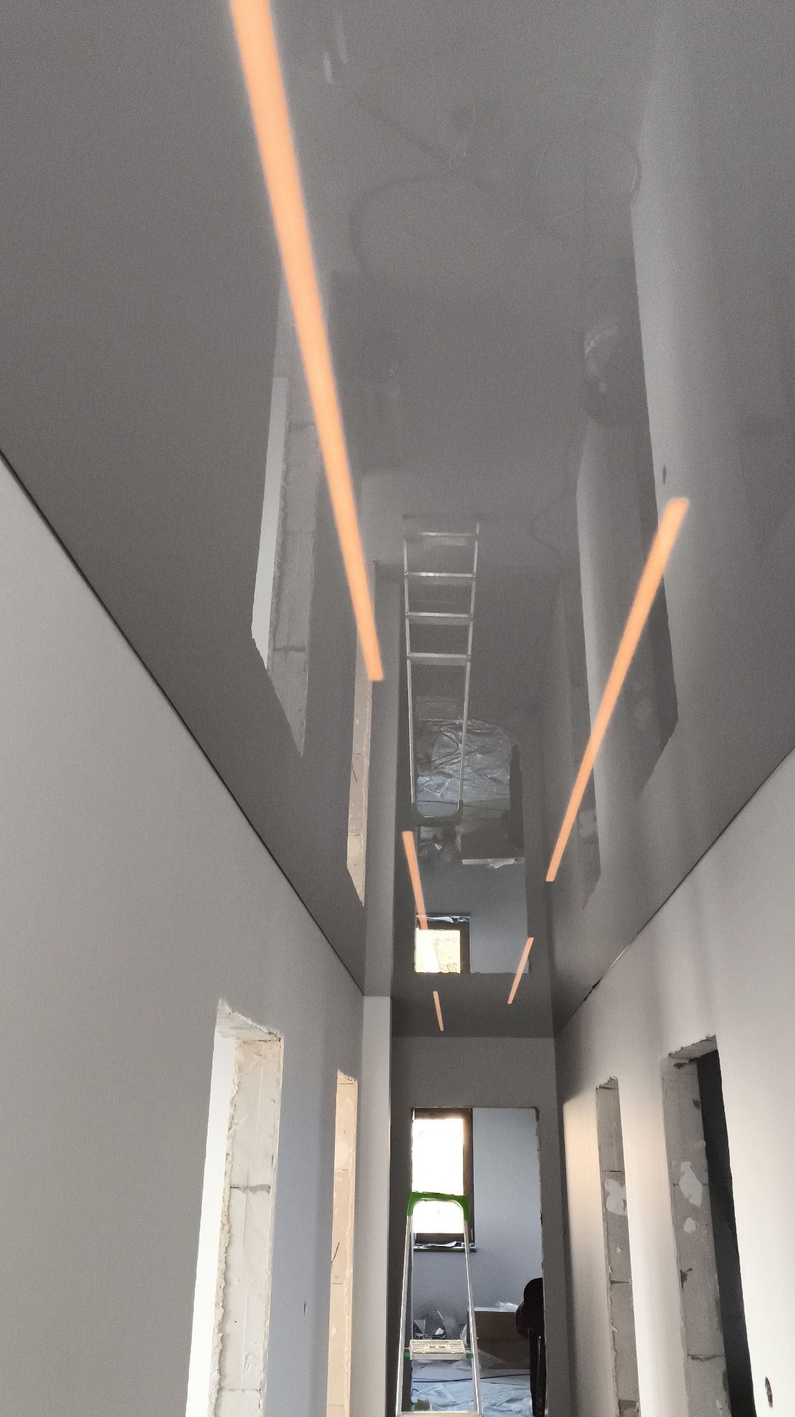 Montaż szarego, lustrzanego sufitu z oświetleniem liniowym w korytarzu. Widoczna drabina i otwory drzwiowe w trakcie wykańczania.