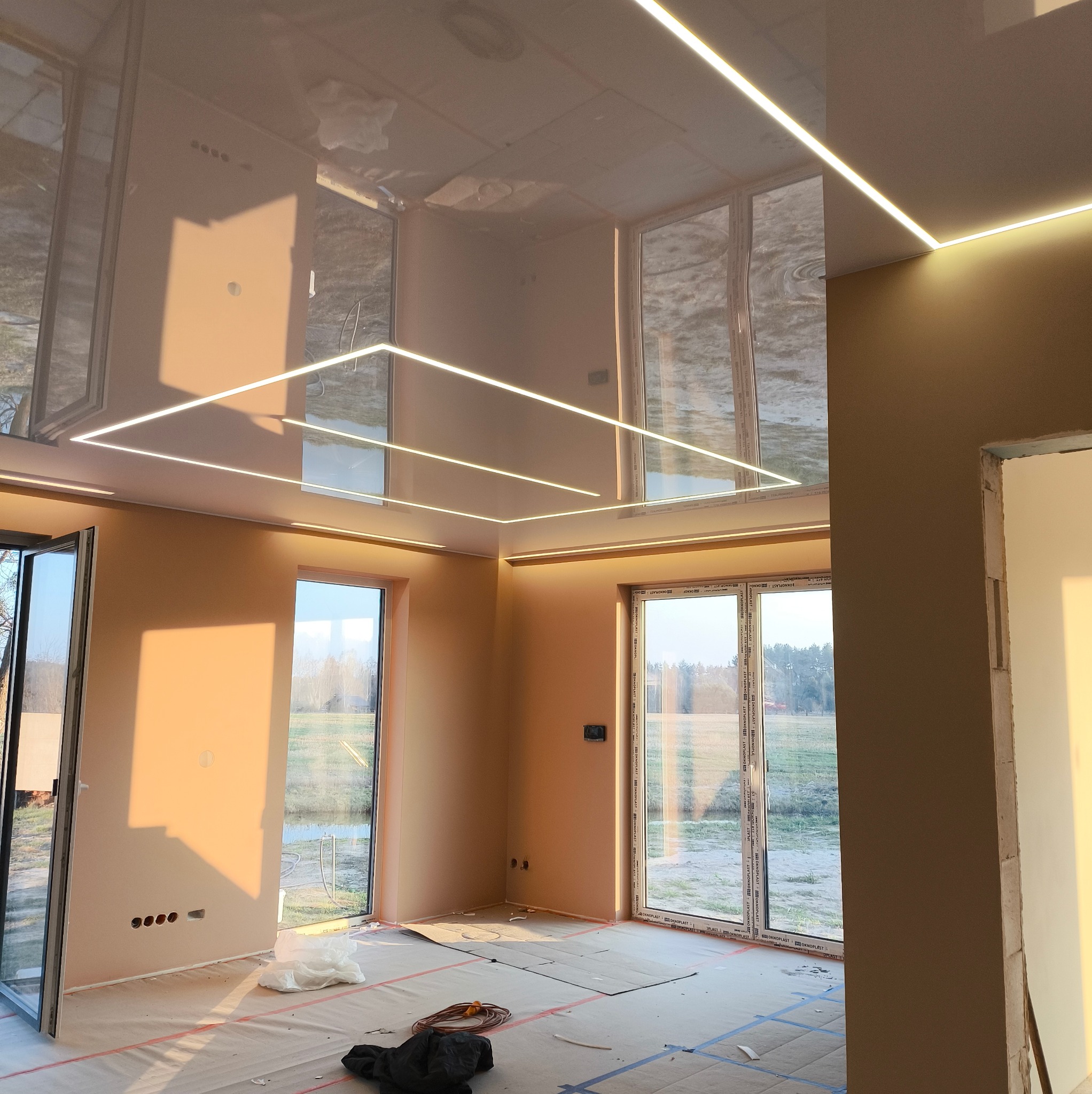 Jasne wnętrze z błyszczącym, podwieszanym sufitem i liniowym oświetleniem LED. Widoczne okna z widokiem na zewnątrz i zabezpieczona podłoga.