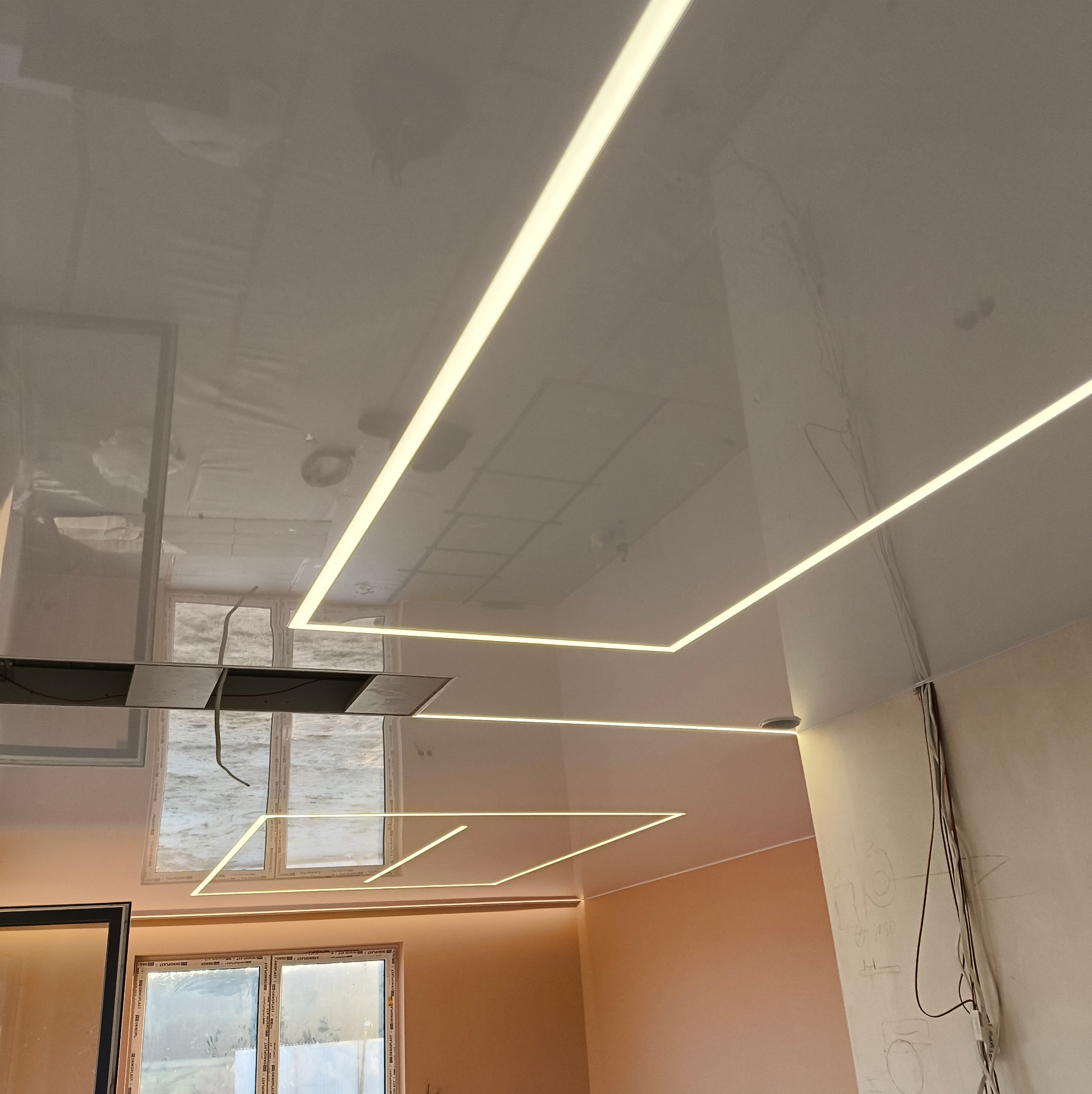 Błyszczący, podwieszany sufit z geometrycznymi liniami LED, odbijający światło z okien. Widoczne kable i notatki na ścianie wskazują na etap instalacji.
