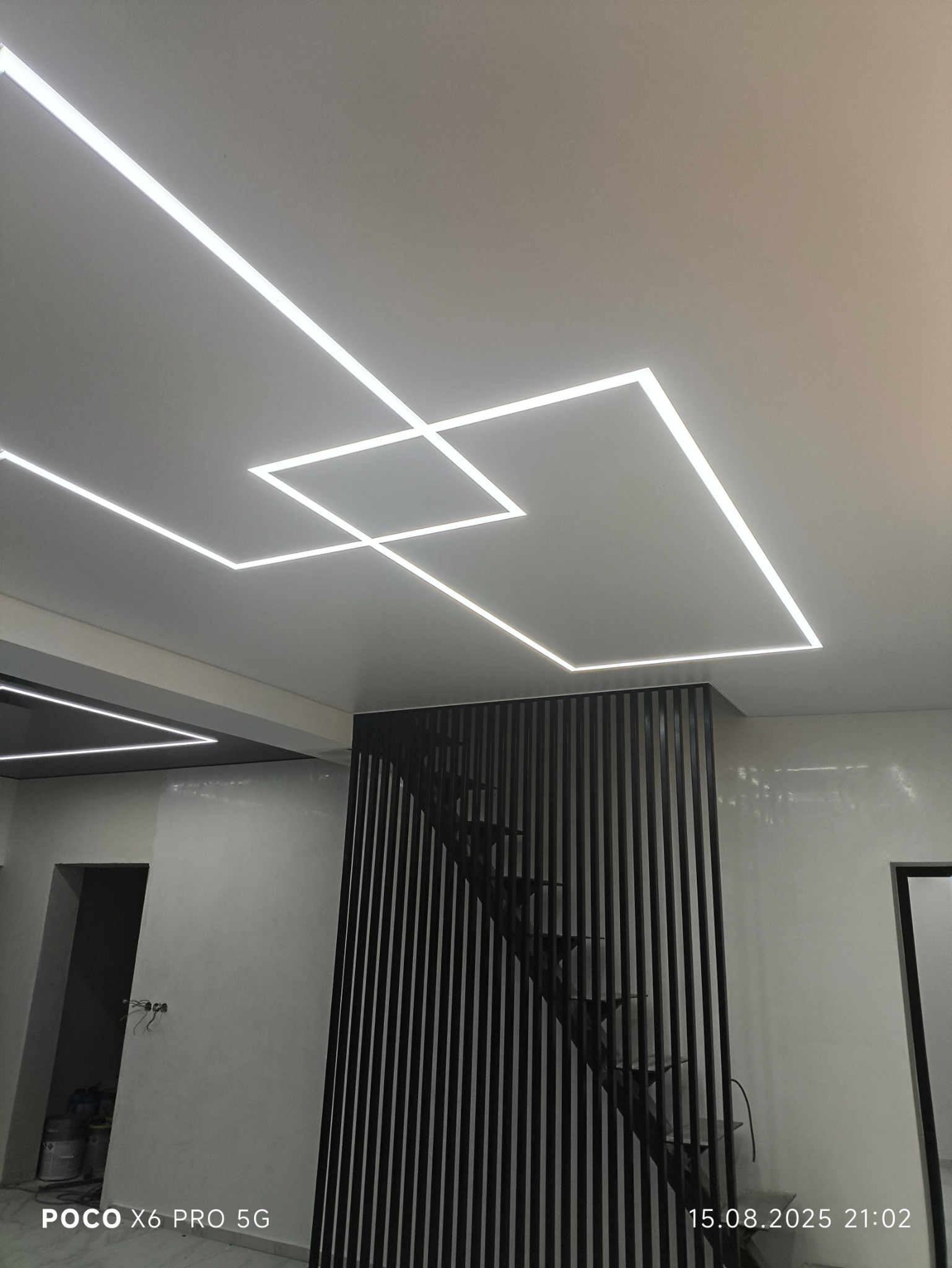 Nowoczesny sufit z geometrycznym wzorem z linii LED, widok z dołu na klatkę schodową z czarną, ażurową balustradą. Minimalistyczny design wnętrza.