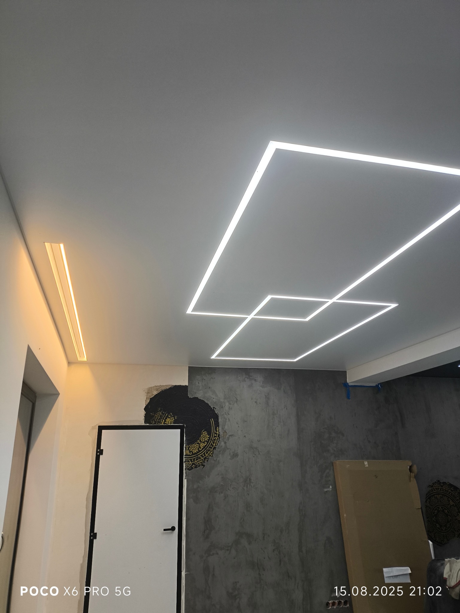 Nowoczesny sufit z oświetleniem LED w kształcie geometrycznym, ciepłe światło podkreśla minimalistyczny design. Widoczne niedokończone wykończenia ścian.