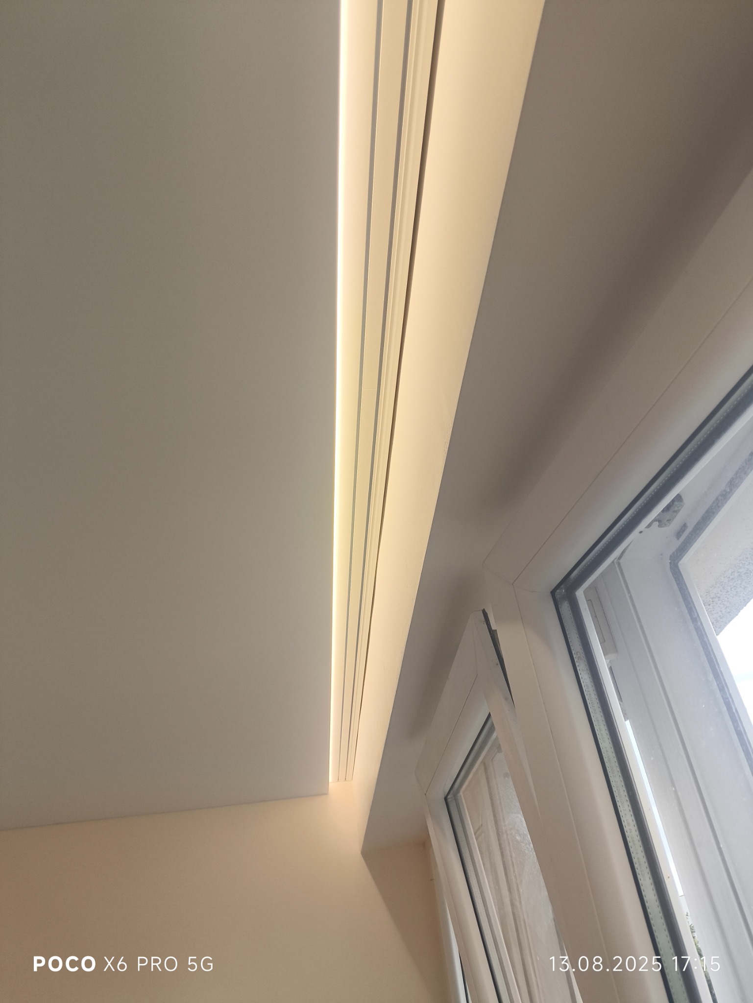 Biały sufit z oświetleniem liniowym LED, widok z dołu, fragment okna. Minimalistyczny design, ciepłe światło. Detal wykończenia wnętrza.