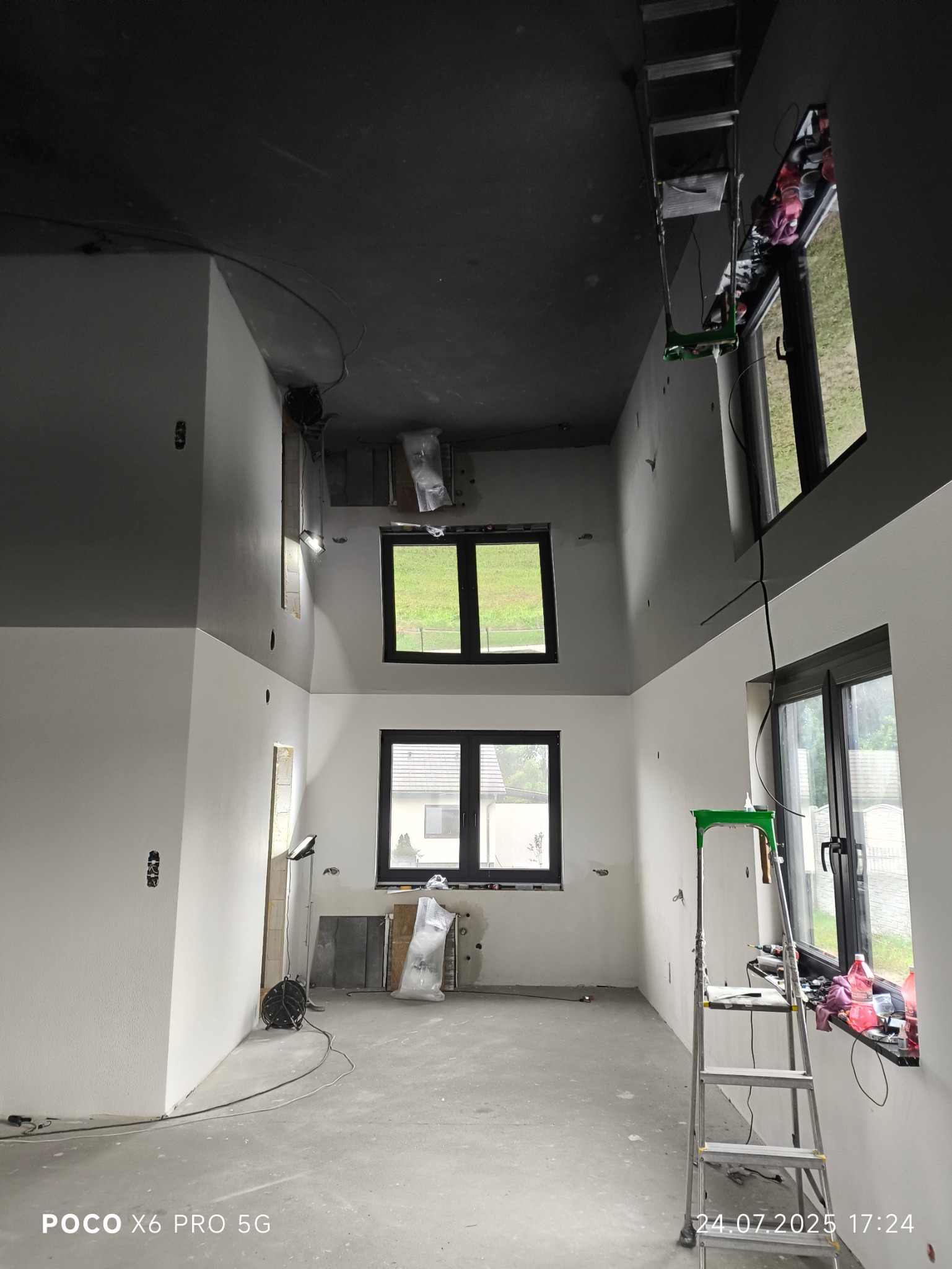 Wysokie wnętrze z czarnym sufitem i białymi ścianami w trakcie montażu. Widoczne okna, drabina i akcesoria montażowe. Surowy stan wykończenia.