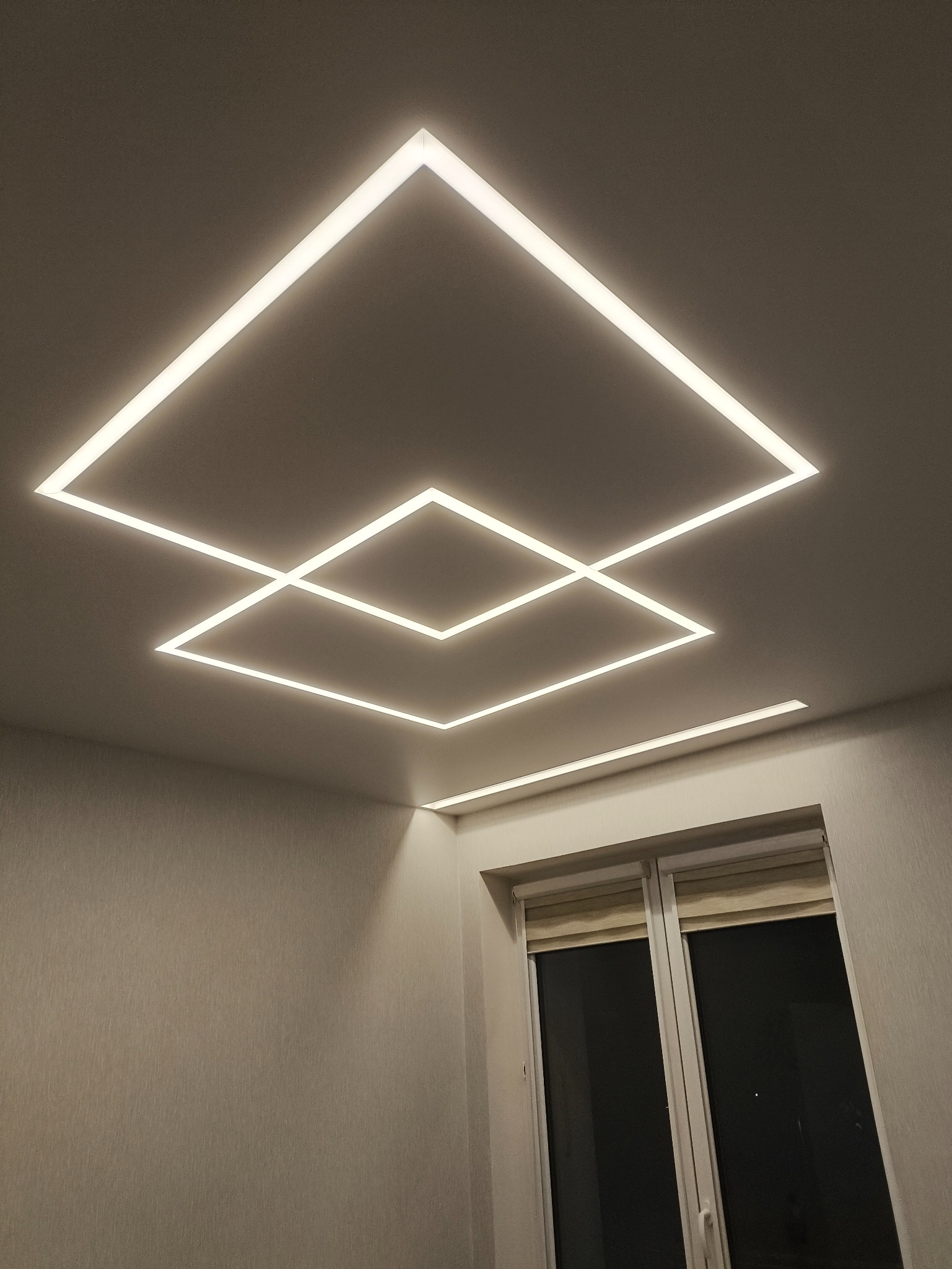 Nowoczesny pokój z geometrycznym oświetleniem LED na suficie, tworzącym kwadratowe wzory. Widoczne okno z roletami i jasne ściany.