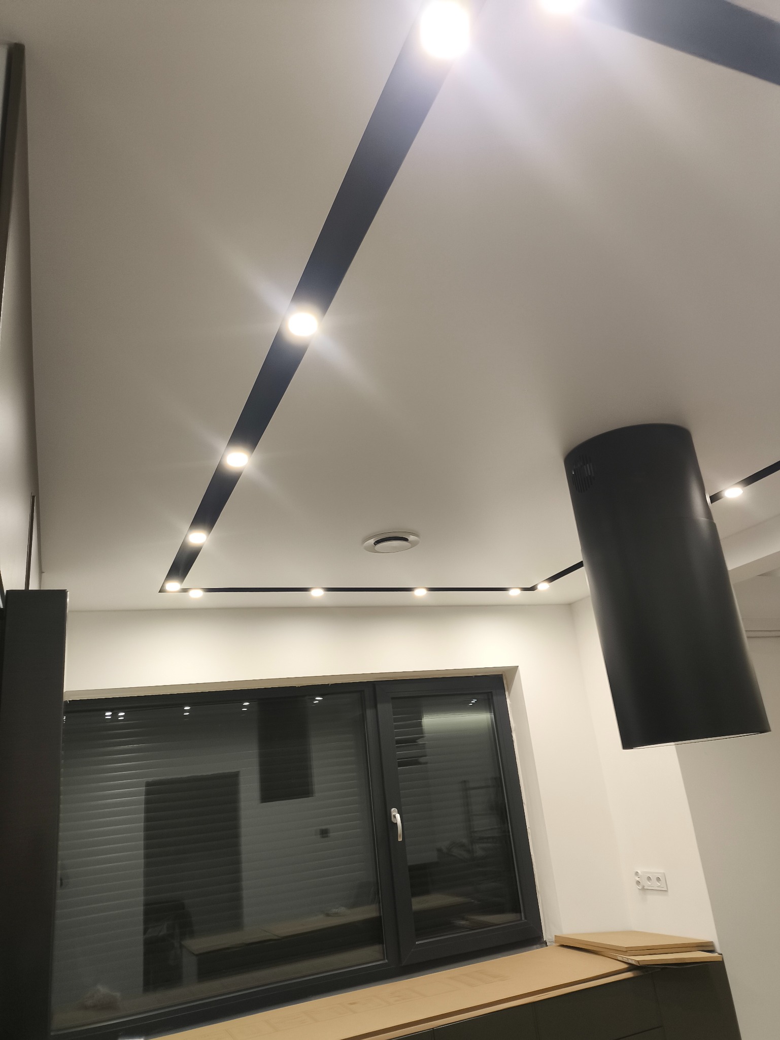 Nowoczesny sufit napinany z czarnymi listwami i punktowym oświetleniem LED w jasnym wnętrzu. Widoczny fragment okna i czarny okap kuchenny.