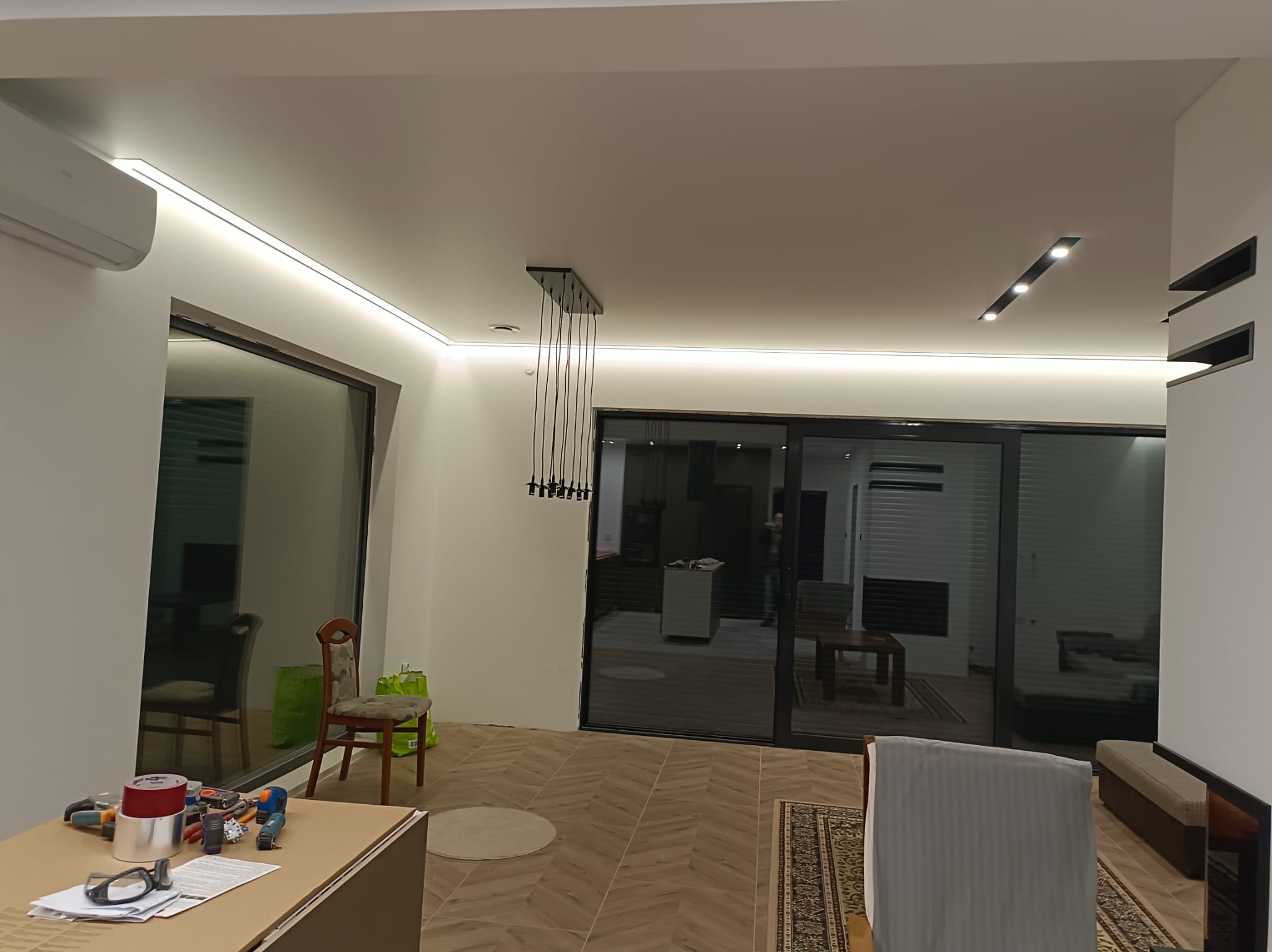 Nowoczesne oświetlenie LED w salonie z podwieszanym sufitem, widoczne narzędzia pracy na pierwszym planie. Minimalistyczny design wnętrza z dużymi oknami.