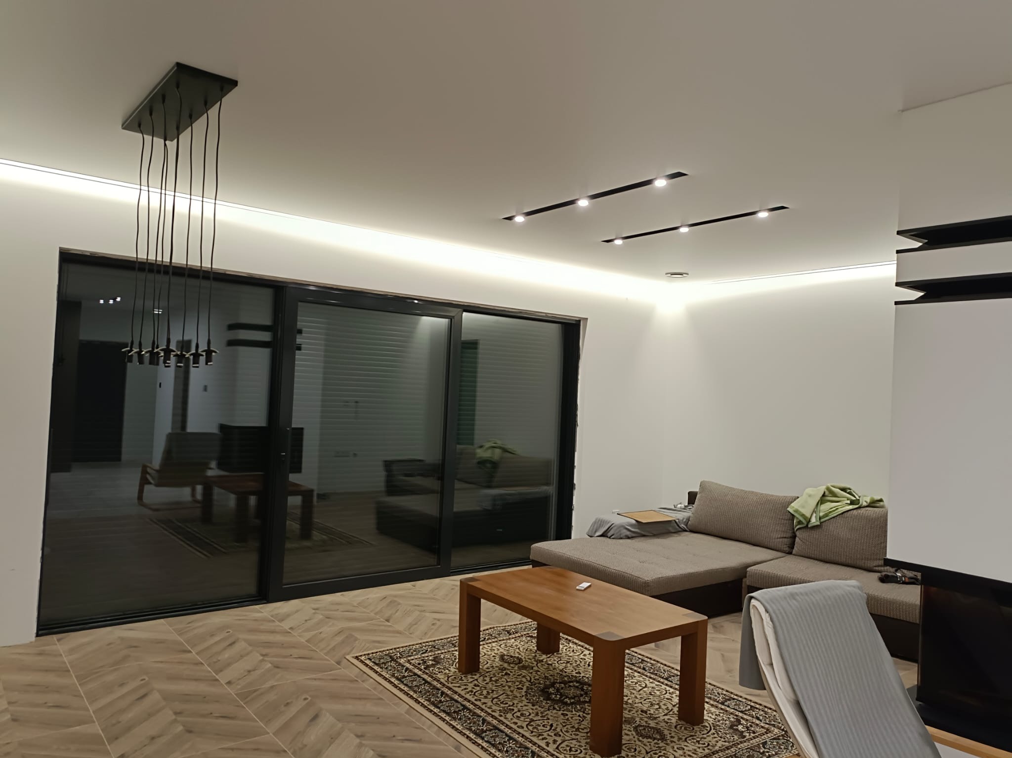 Nowoczesny salon z oświetleniem sufitowym: czarne listwy LED i designerska lampa wisząca. Parkiet w jodełkę, szara sofa i stolik kawowy na wzorzystym dywanie.