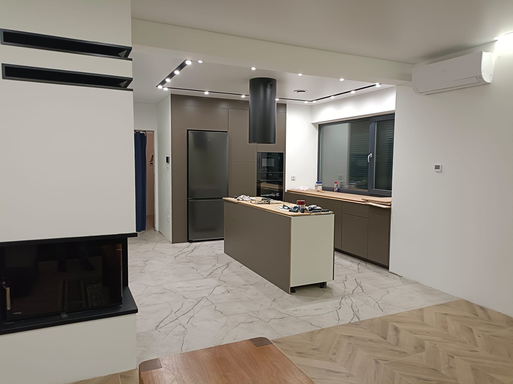 Nowoczesna kuchnia z wyspą i marmurową podłogą, oświetlenie punktowe, minimalistyczny design, widok na salon z kominkiem. Ukończone wnętrze.