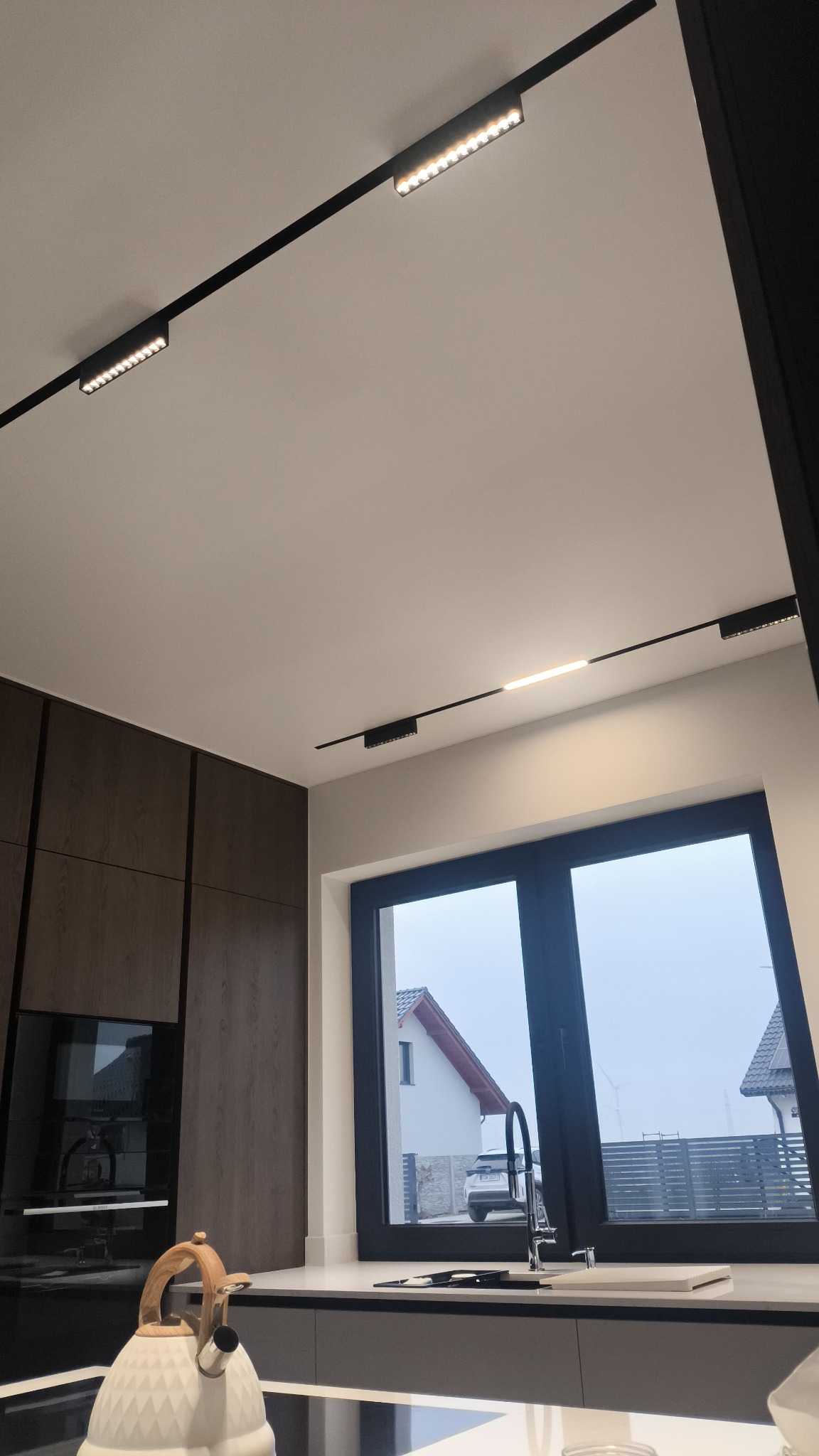 Nowoczesna kuchnia z oknem: czarne szyny oświetleniowe LED na białym suficie, minimalistyczny design, widok na dom zza okna.