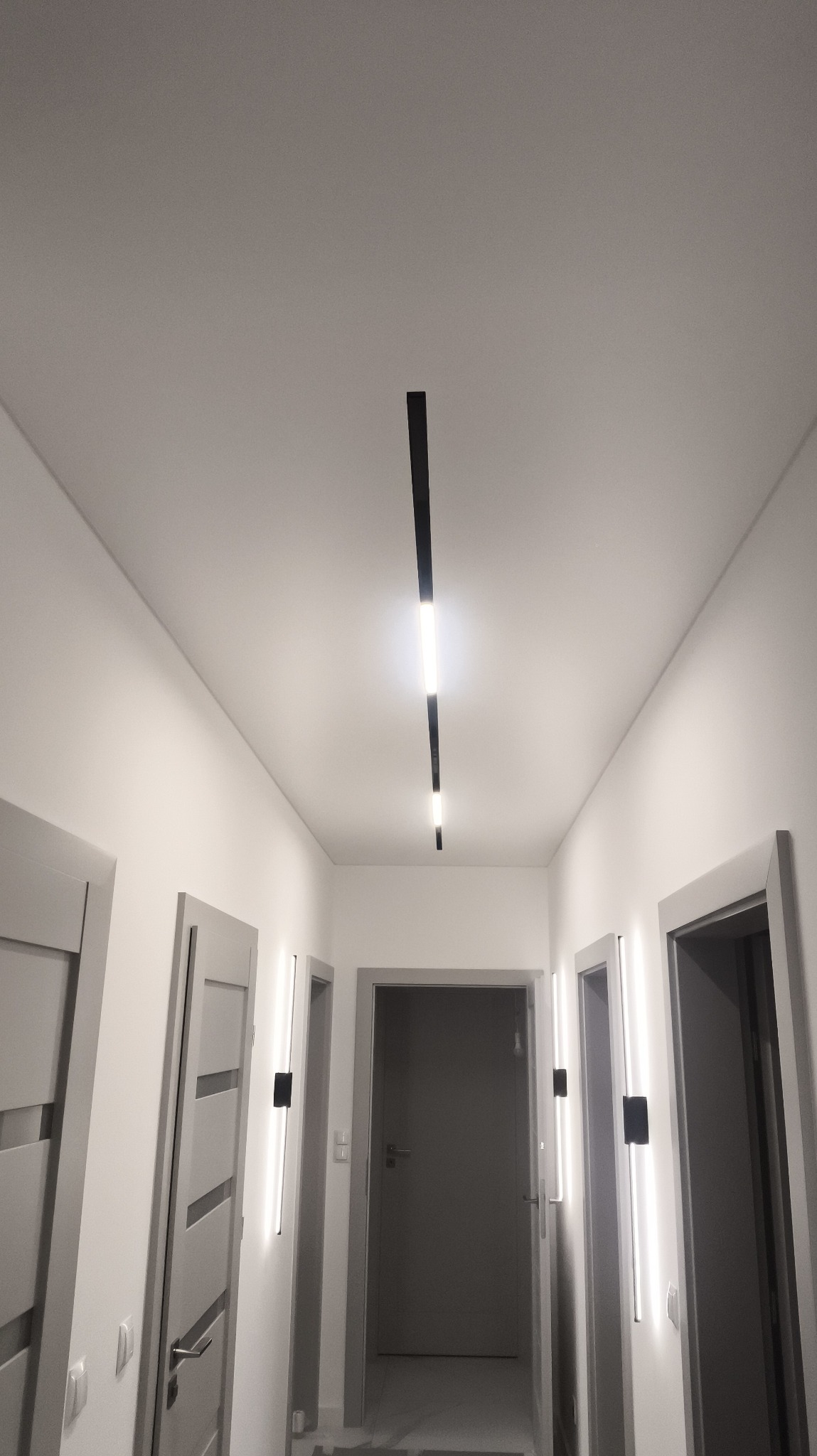 Nowoczesny korytarz z białym sufitem i liniowym oświetleniem LED. Szare drzwi z ościeżnicami i pionowe lampy ścienne dodają elegancji.