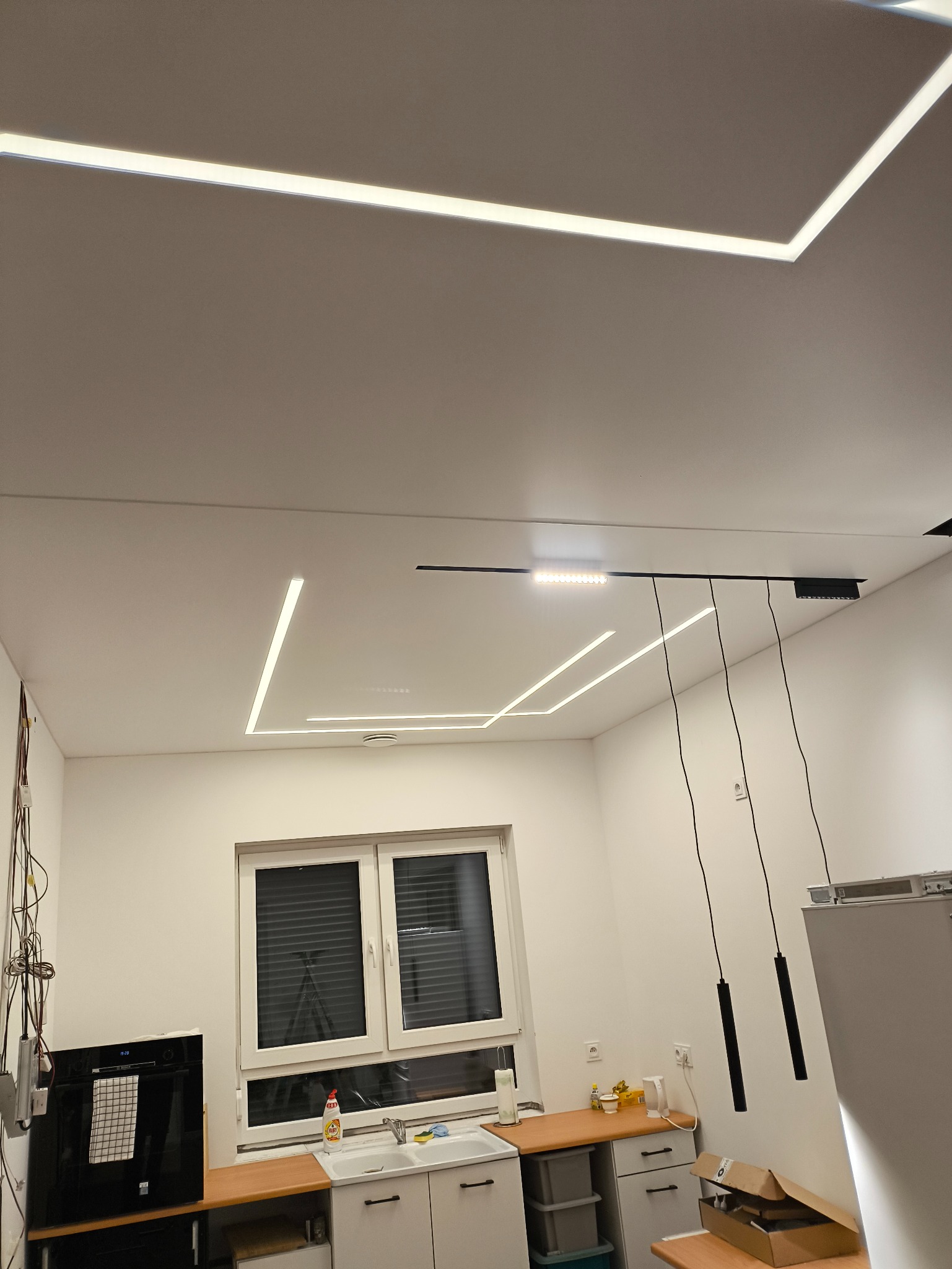 Nowoczesny sufit z liniowym oświetleniem LED w kształcie prostokątów i wisząca lampa nad blatem kuchennym. Minimalistyczny design, białe ściany, okno z żaluzjami.
