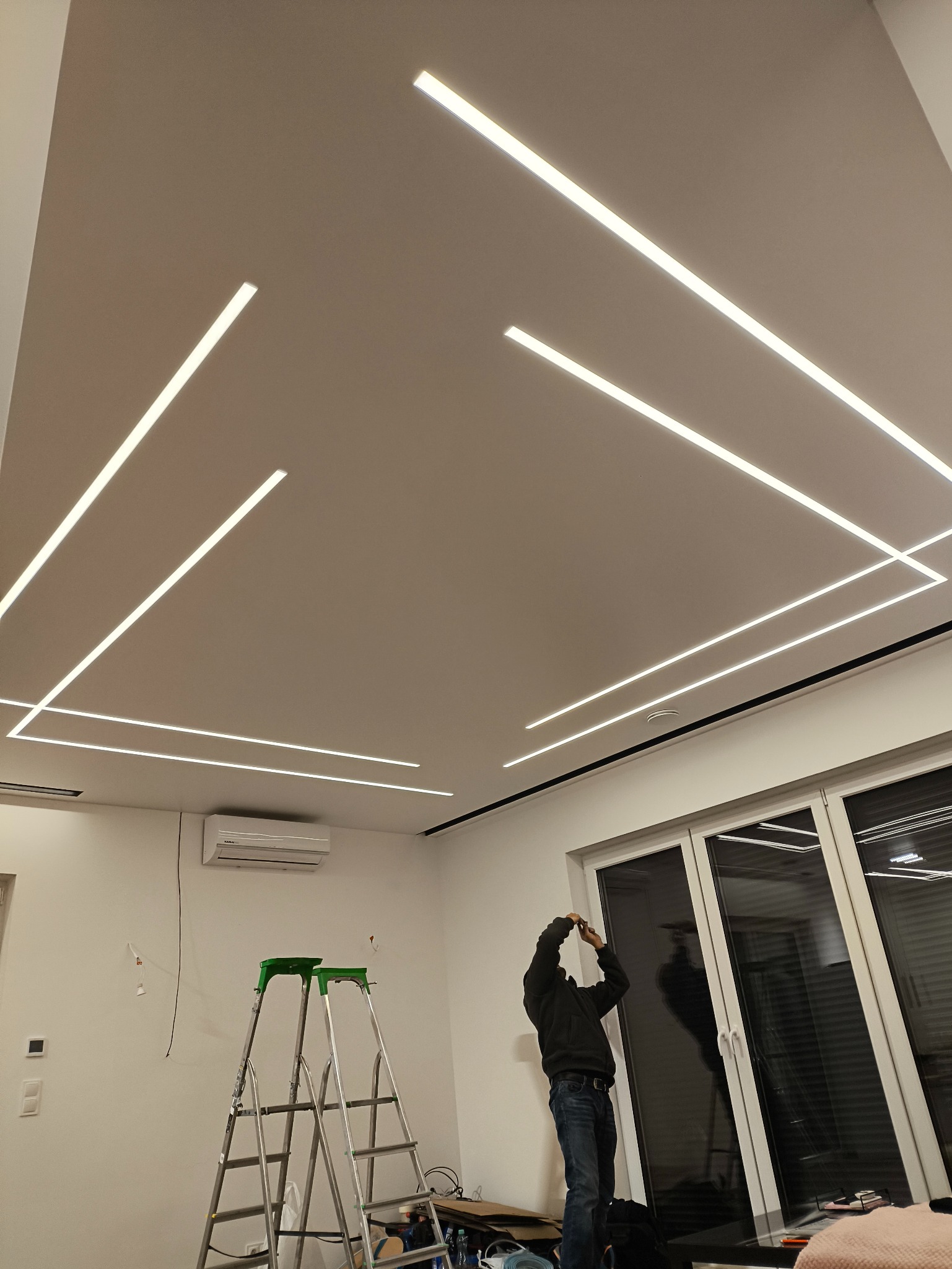 Montaż liniowego oświetlenia LED na suficie podwieszanym. Widoczna drabina i instalator podczas pracy. Nowoczesne wnętrze z klimatyzacją i dużymi oknami.