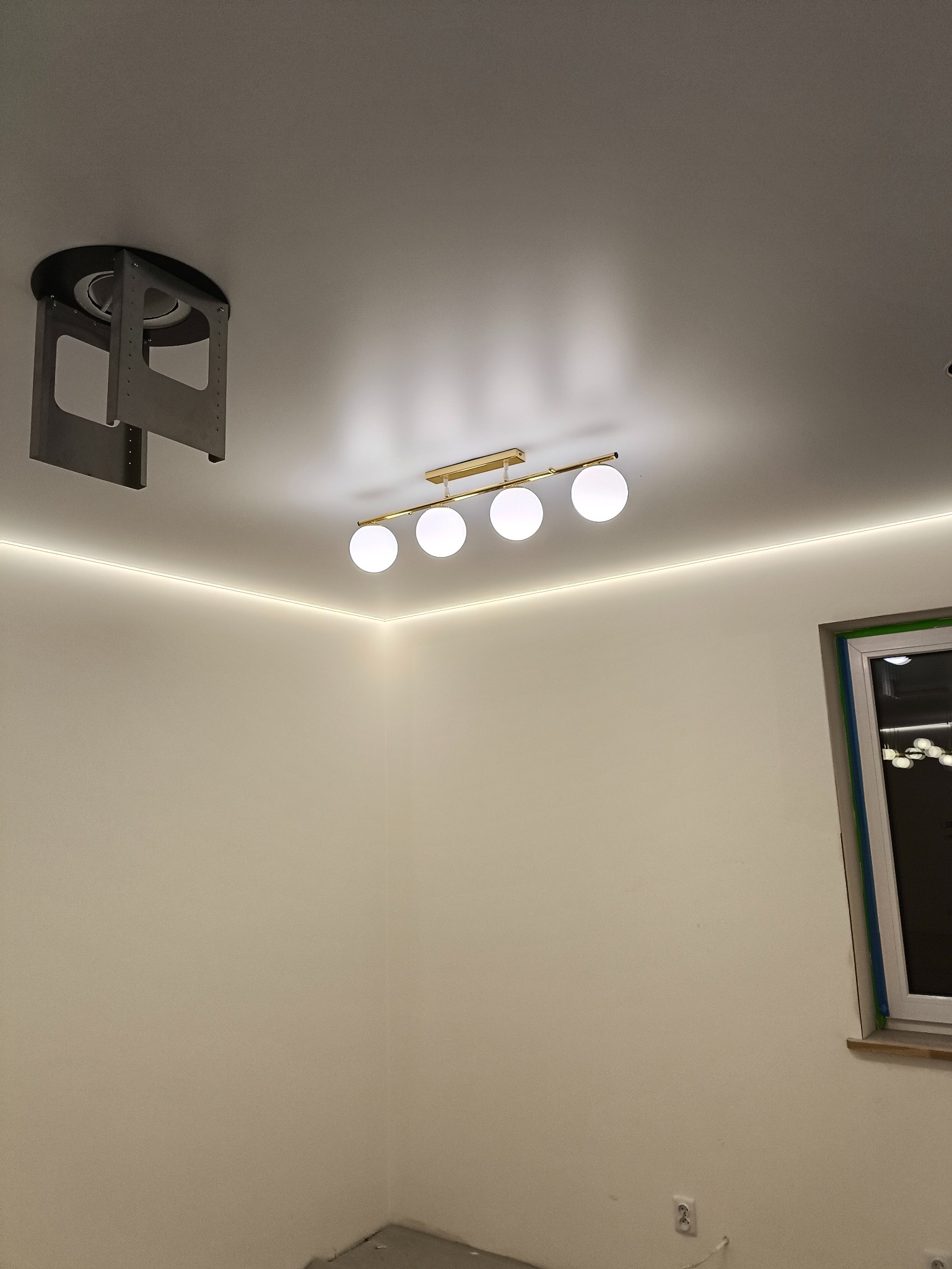 Nowoczesne oświetlenie sufitowe z czterema kulistymi lampami i listwą LED wzdłuż ściany. Widoczny element montażowy. Świeżo wykończone wnętrze.