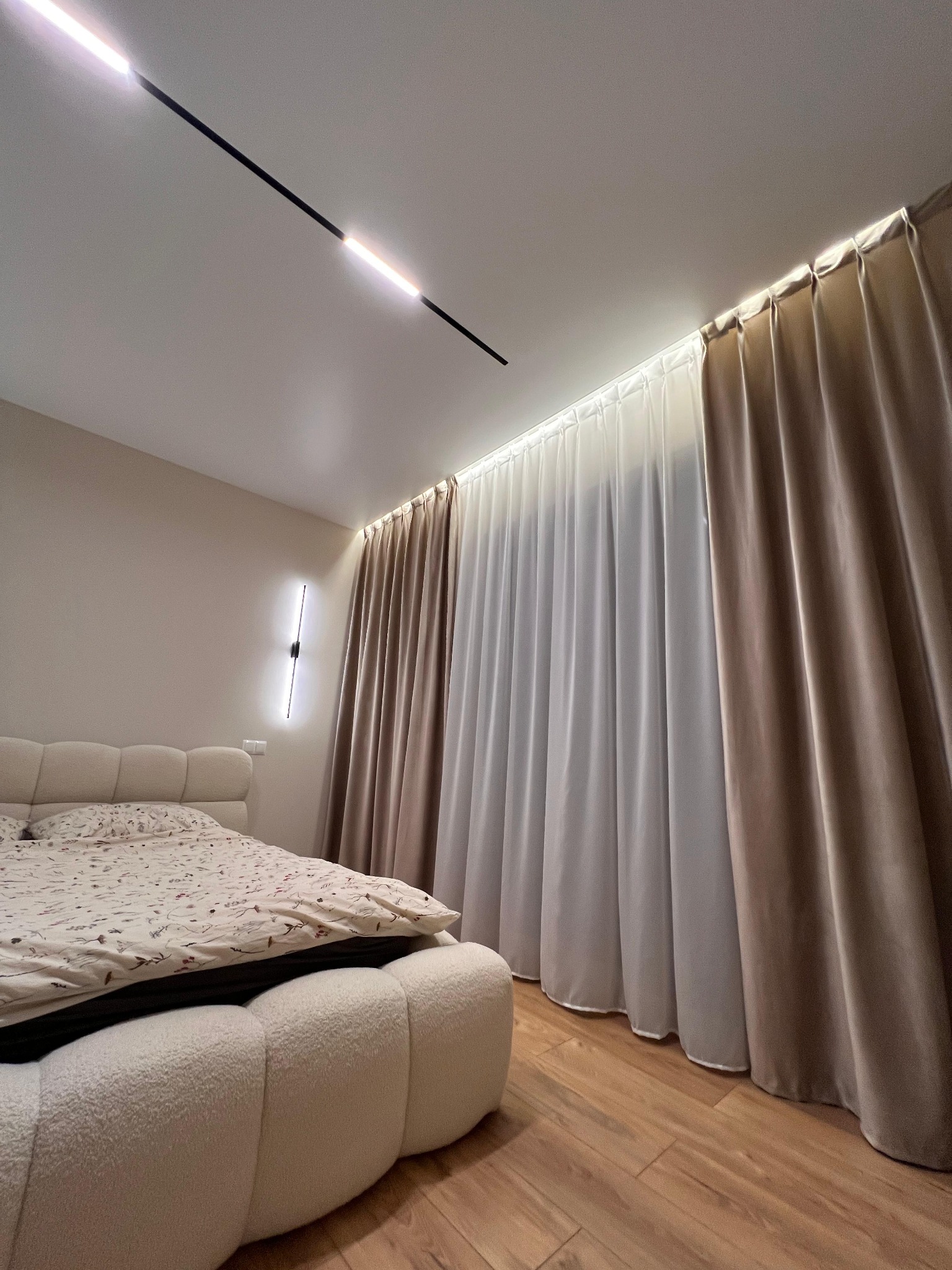 Nowoczesna sypialnia z podwieszanym oświetleniem sufitowym, beżowymi zasłonami i dekoracyjną lampą ścienną. Widok na łóżko i drewnianą podłogę.
