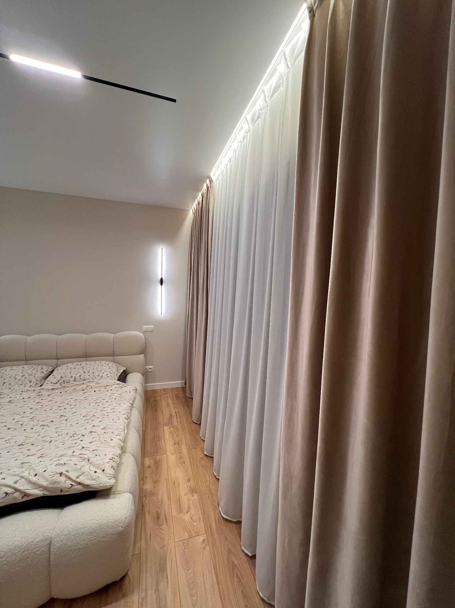 Nowoczesna sypialnia z beżowymi zasłonami i podświetlanym sufitem. Minimalistyczna lampa ścienna i drewniana podłoga dodają elegancji wnętrzu.