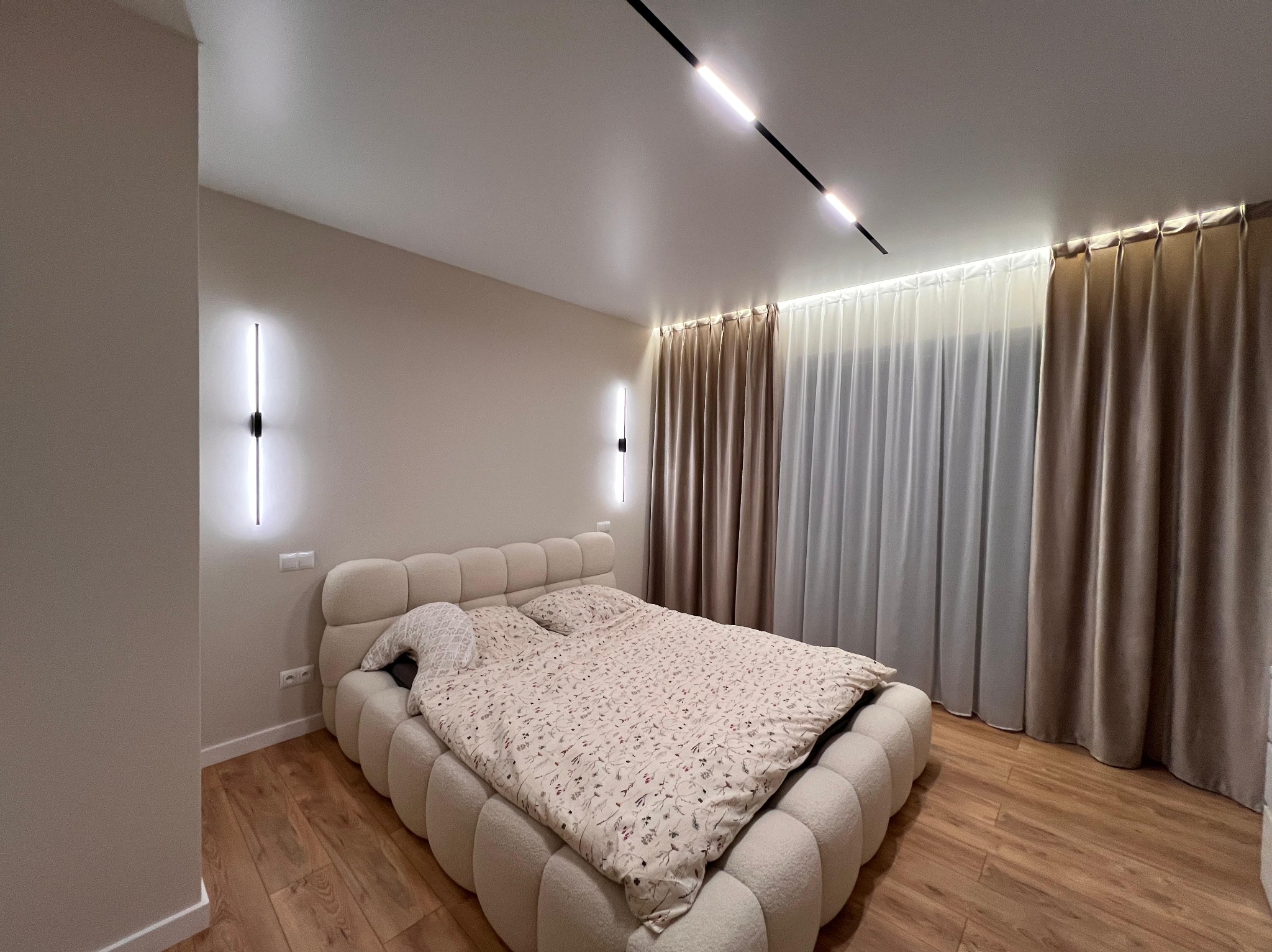 Nowoczesna sypialnia z podwieszanym oświetleniem szynowym i minimalistycznymi kinkietami ściennymi, beżowe ściany, drewniana podłoga i pluszowe łóżko.