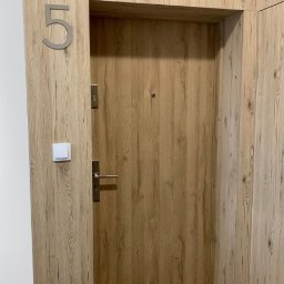 Piętro i Parter - Drewniana okleina drzwi wejściowych z numerem '5', srebrna klamka i wizjer. W tle widoczny fragment ściany i podłogi. Całość utrzymana w jasnej kolorystyce drewna.