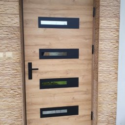 Piętro i Parter - Nowoczesne drzwi zewnętrzne w odcieniach drewna z czarnymi, prostokątnymi przeszkleniami, zamontowane w elewacji wykończonej kamieniem dekoracyjnym.