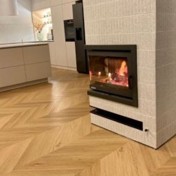 Piętro i Parter - Jasny parkiet ułożony w jodełkę francuską, uzupełniony o nowoczesny kominek z obudową z płytek. W tle widoczna zabudowa kuchenna w neutralnych barwach.