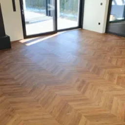 Nowoczesne wnętrze z podłogą ułożoną w jodełkę z paneli o ciepłym, brązowym odcieniu. Widoczne duże okna przesuwne z czarnymi ramami i minimalistyczne ściany.