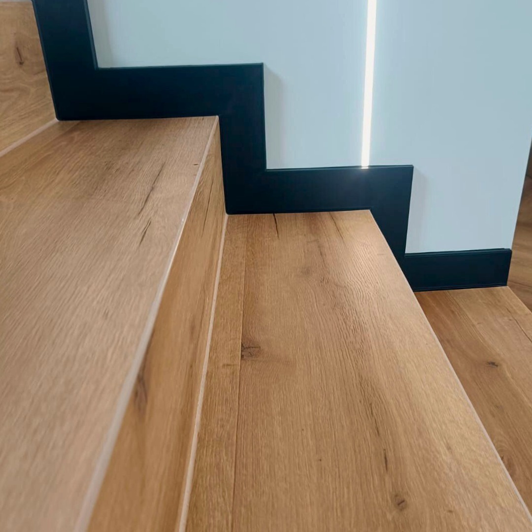 Detale wykończenia schodów z drewnopodobnych paneli, z czarnymi listwami przypodłogowymi i oświetleniem LED przy ścianie. Ujęcie z bliska, podkreślające fakturę materiału.